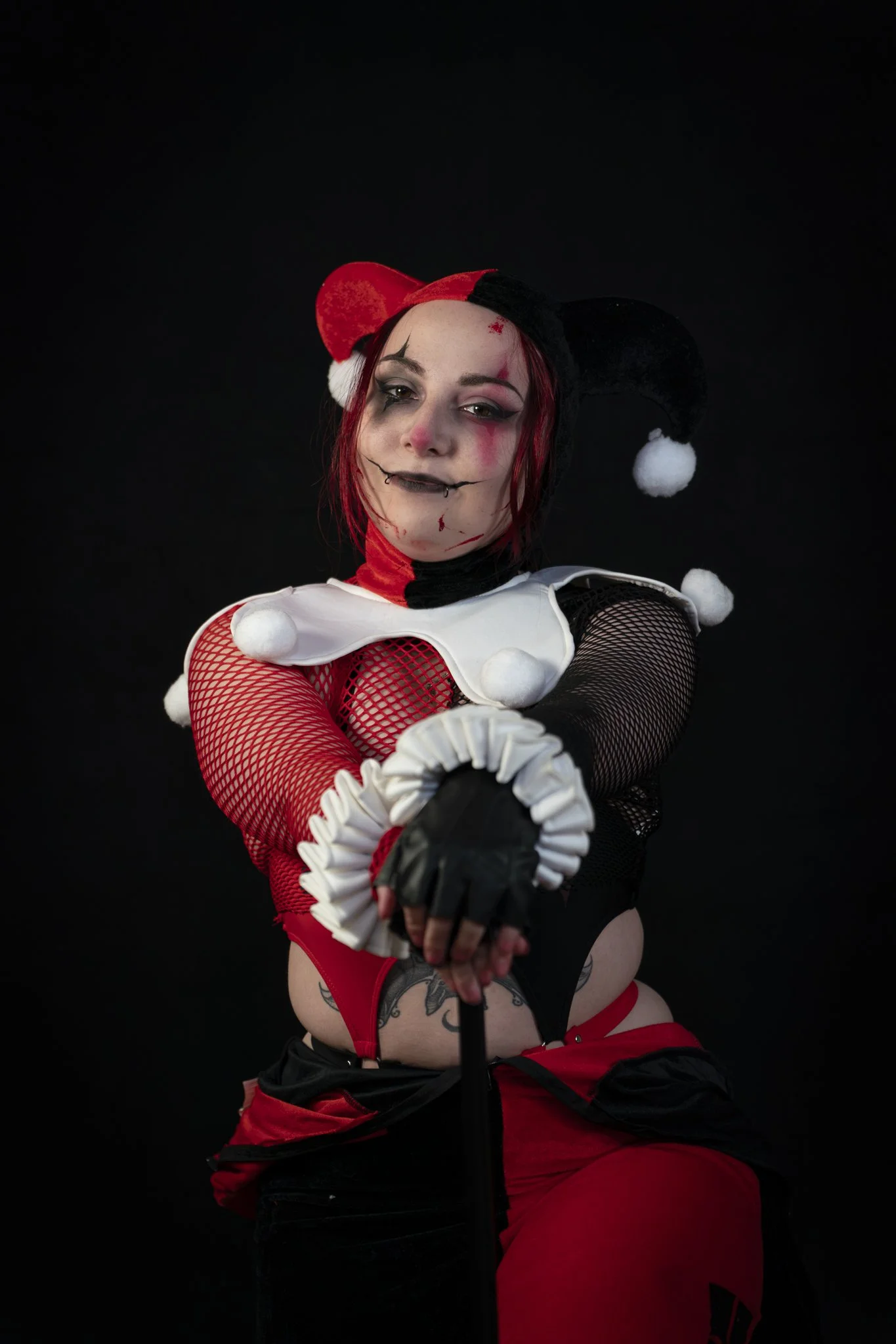 A person in a Harley Quinn-inspired costume with black, red, and white colors, fishnet gloves, and a jester hat, holding a microphone against a black background.