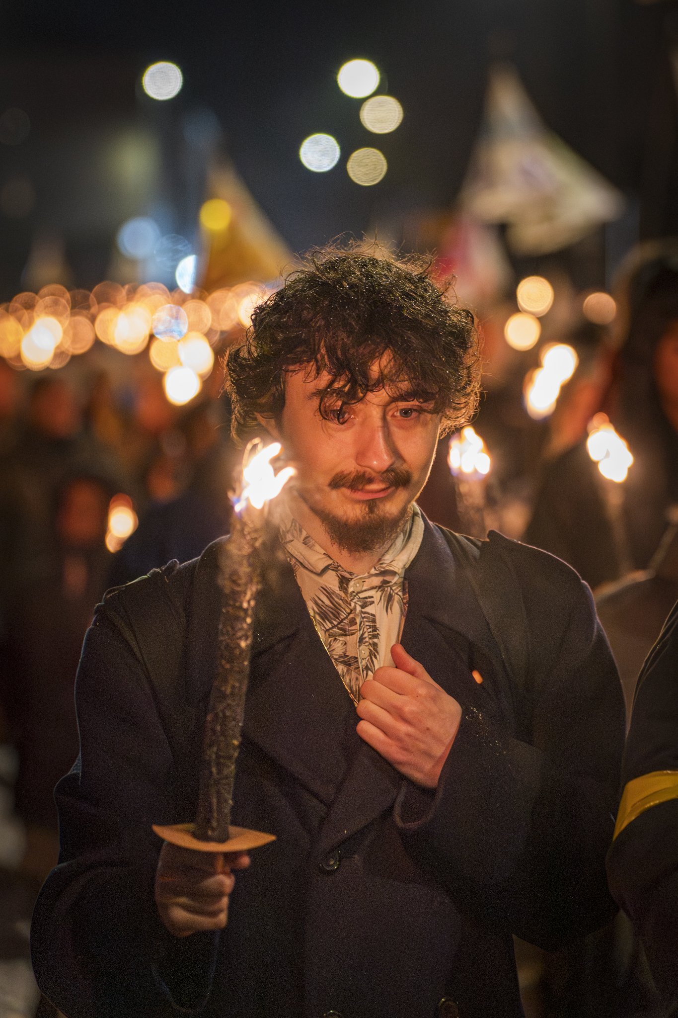A man with dark curly hair, a mustache, and a beard holding a lit torch at night during a celebration or festival, with blurred colorful lights and tents in the background.