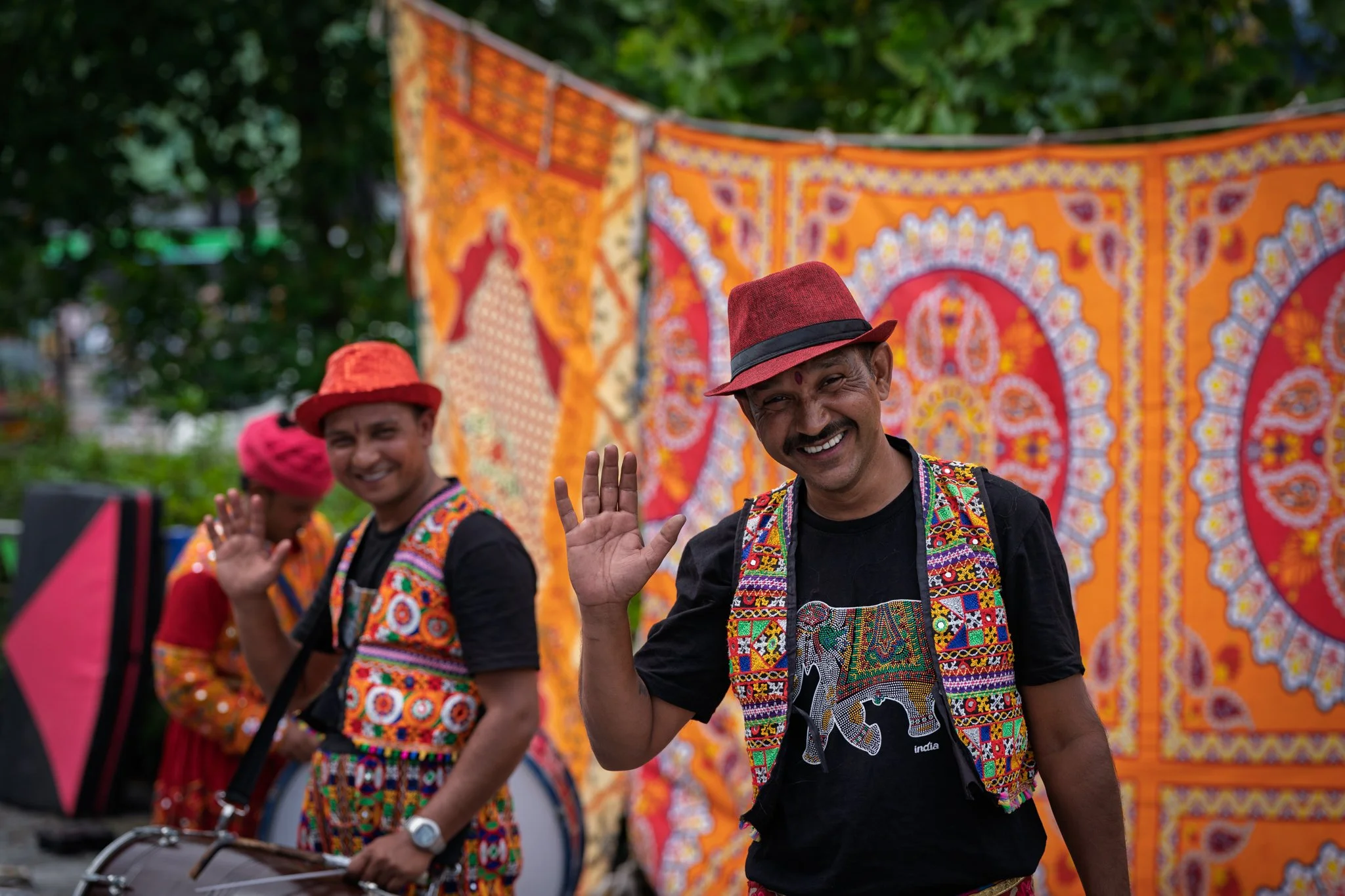 Three men in traditional colorful embroidered Indian attire waving and smiling at a cultural event. Behind them is a vibrant orange decorated backdrop with intricate patterns.
