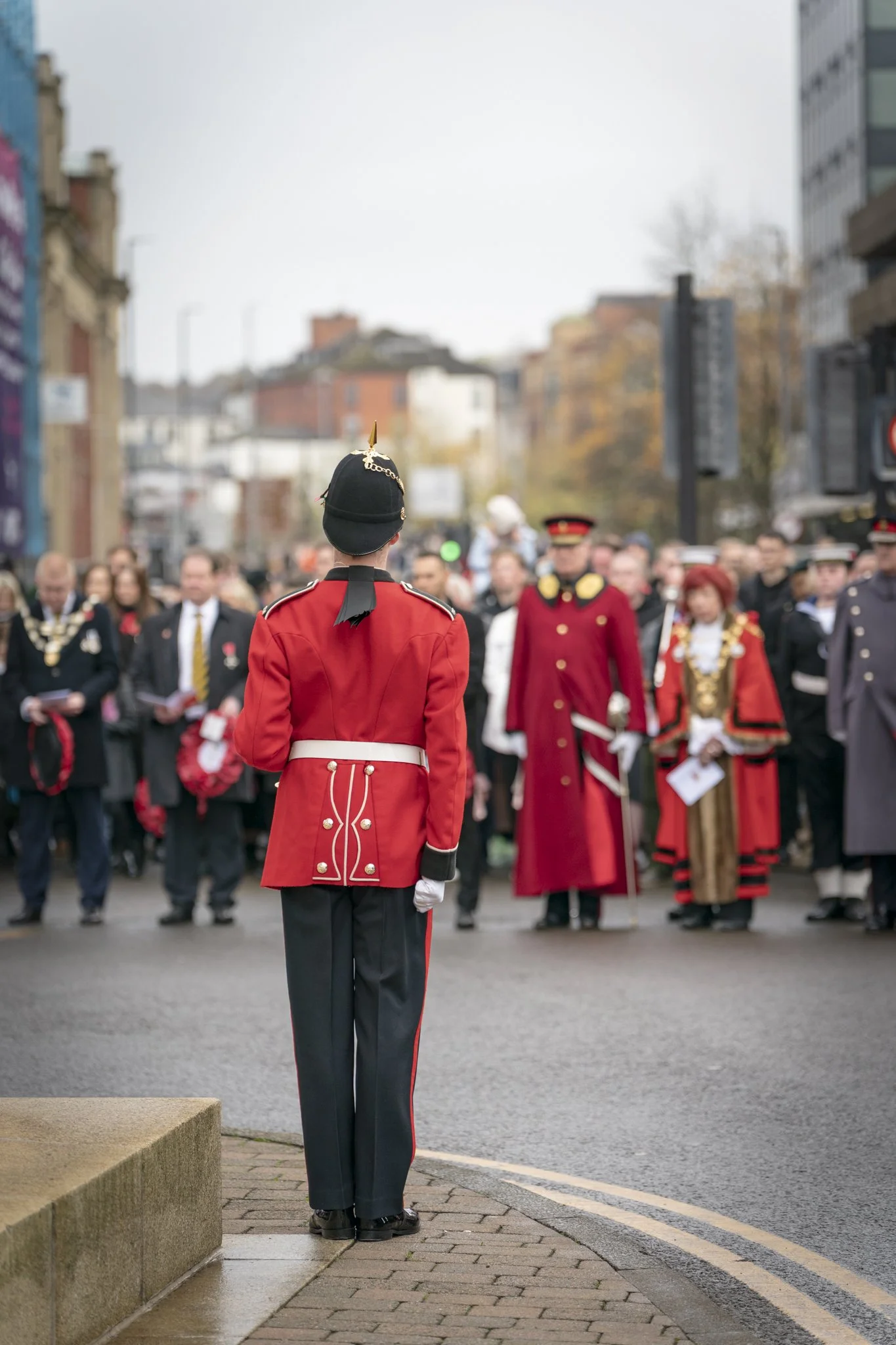 A parade marshal in a red uniform faces a crowd of people dressed in formal and traditional attire on a city street.
