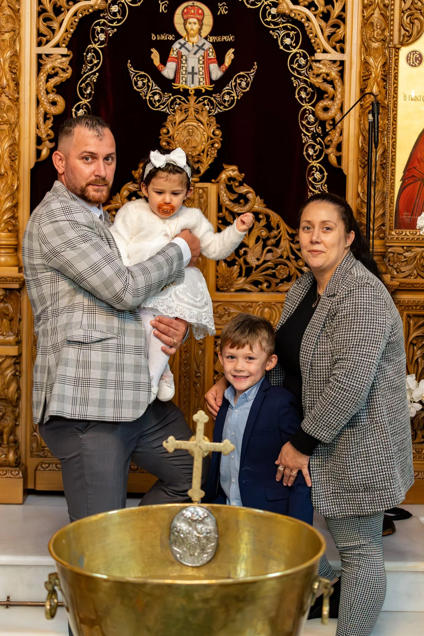 A family dressed in formal attire posing inside a church after a baptism ceremony. The father is holding a young girl with a pacifier in her mouth, and a woman is standing next to a young boy. The background features ornate wood carvings and religiou