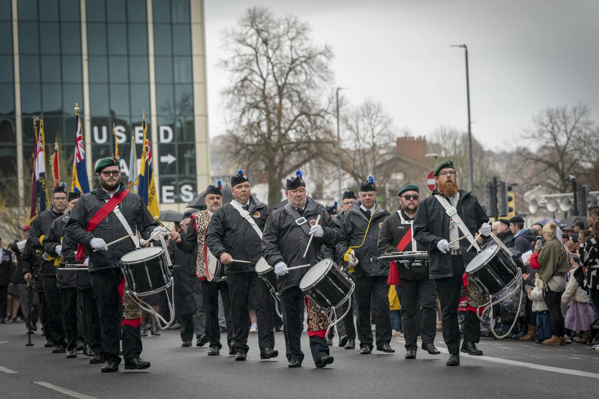 A marching band performing outdoors in front of a crowd, with flags and a modern glass building with the words 'USED' and an arrow in the background. The band members wear black uniforms with green berets, some have sunglasses, and many are playing d