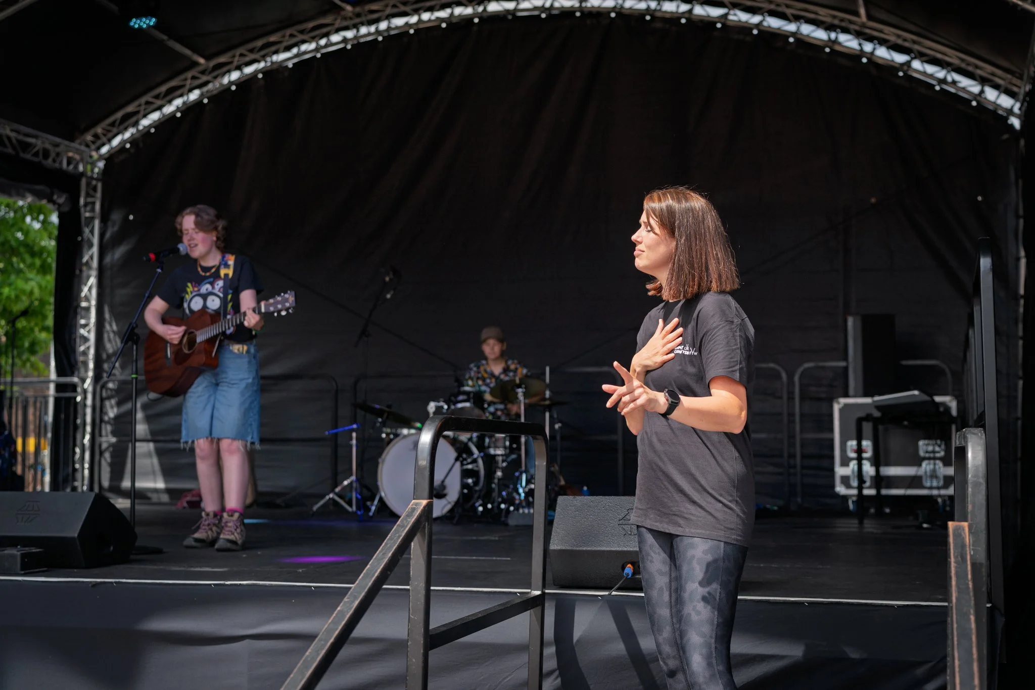 A woman singing with her hand on her chest on stage, while a young girl plays guitar and sings in the background at an outdoor concert.