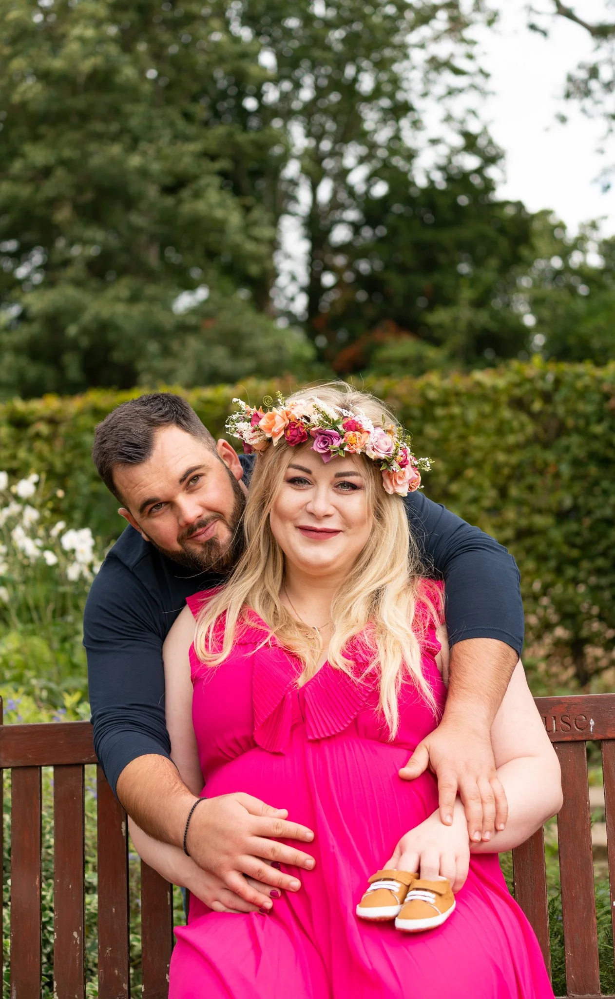 A couple sitting on a wooden bench outdoors, with the woman in a bright pink dress and a floral crown, and the man embracing her from behind.