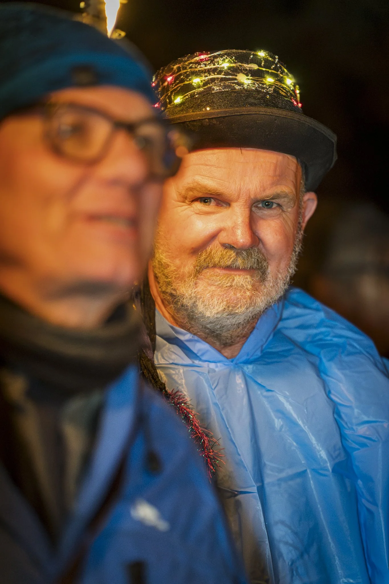 A man with a beard wearing a black hat decorated with colorful string lights, and a blue jacket. Another person in the foreground wearing glasses, a blue beanie, and a dark jacket. The background is dark, suggesting this is taken at night.