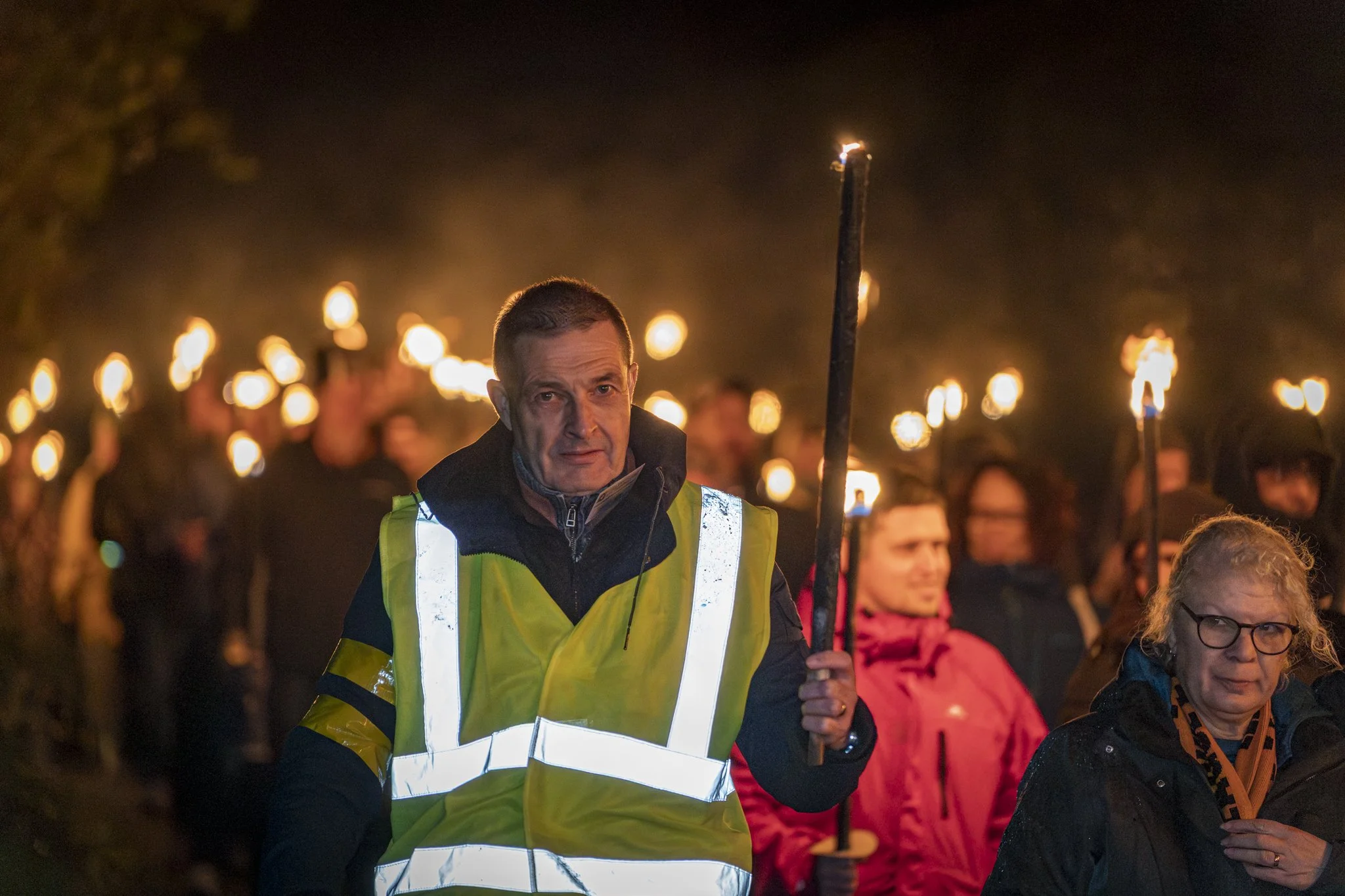 Man in a yellow reflective vest holding a torch during a nighttime event with a crowd holding torches.