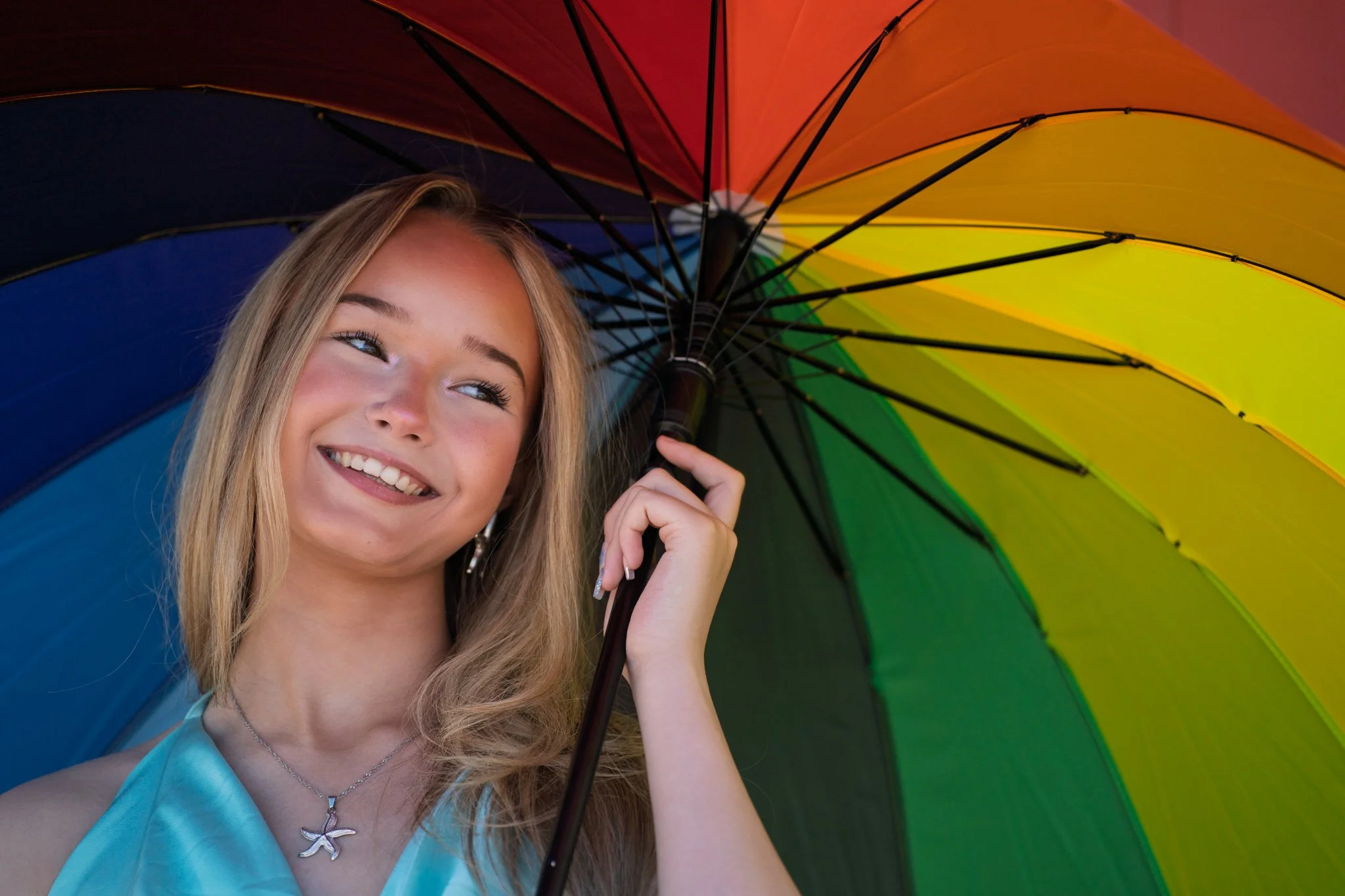 A young woman with long blonde hair smiling and holding a rainbow-colored umbrella.