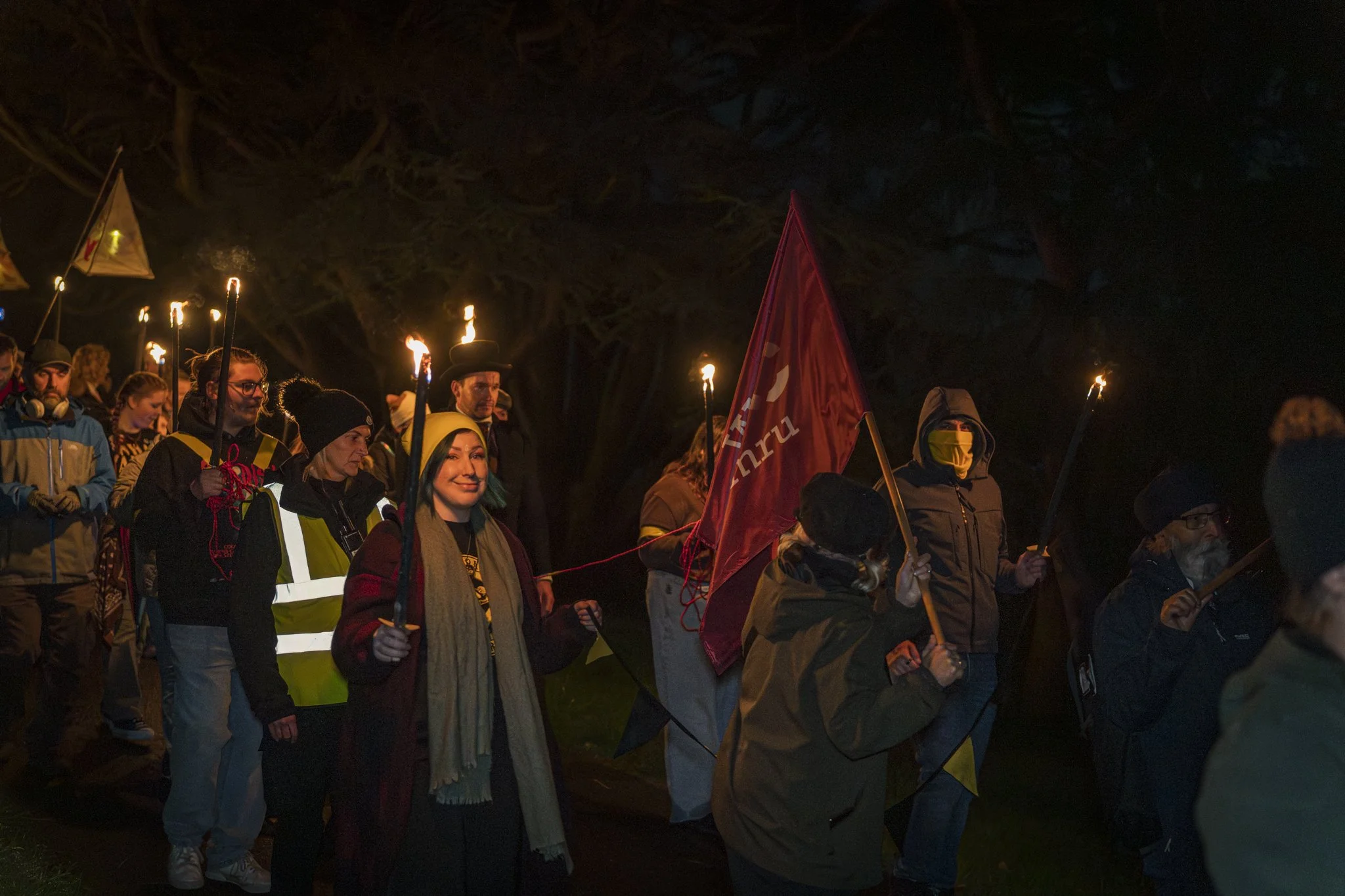 A group of people participating in a nighttime protest or march, holding torches and flags, some wearing reflective vests and face coverings.