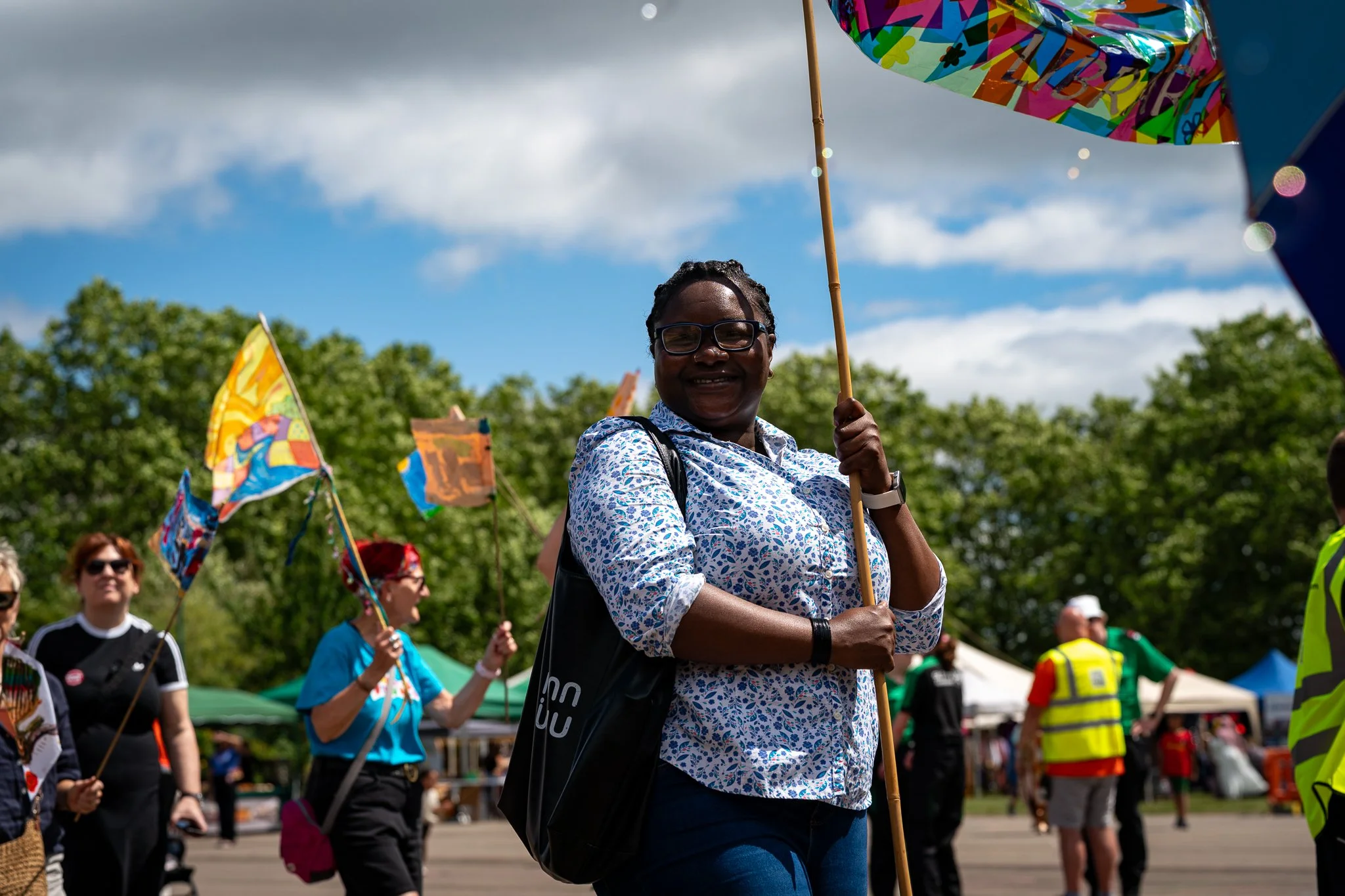 A woman smiling and holding a colorful umbrella at an outdoor event or rally, surrounded by other people. The sky is partly cloudy with trees in the background.