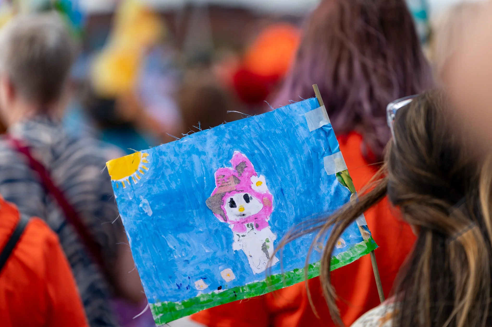 Children holding a painted art piece with a pink character wearing a hat, blue background, and flowers, at an outdoor event.