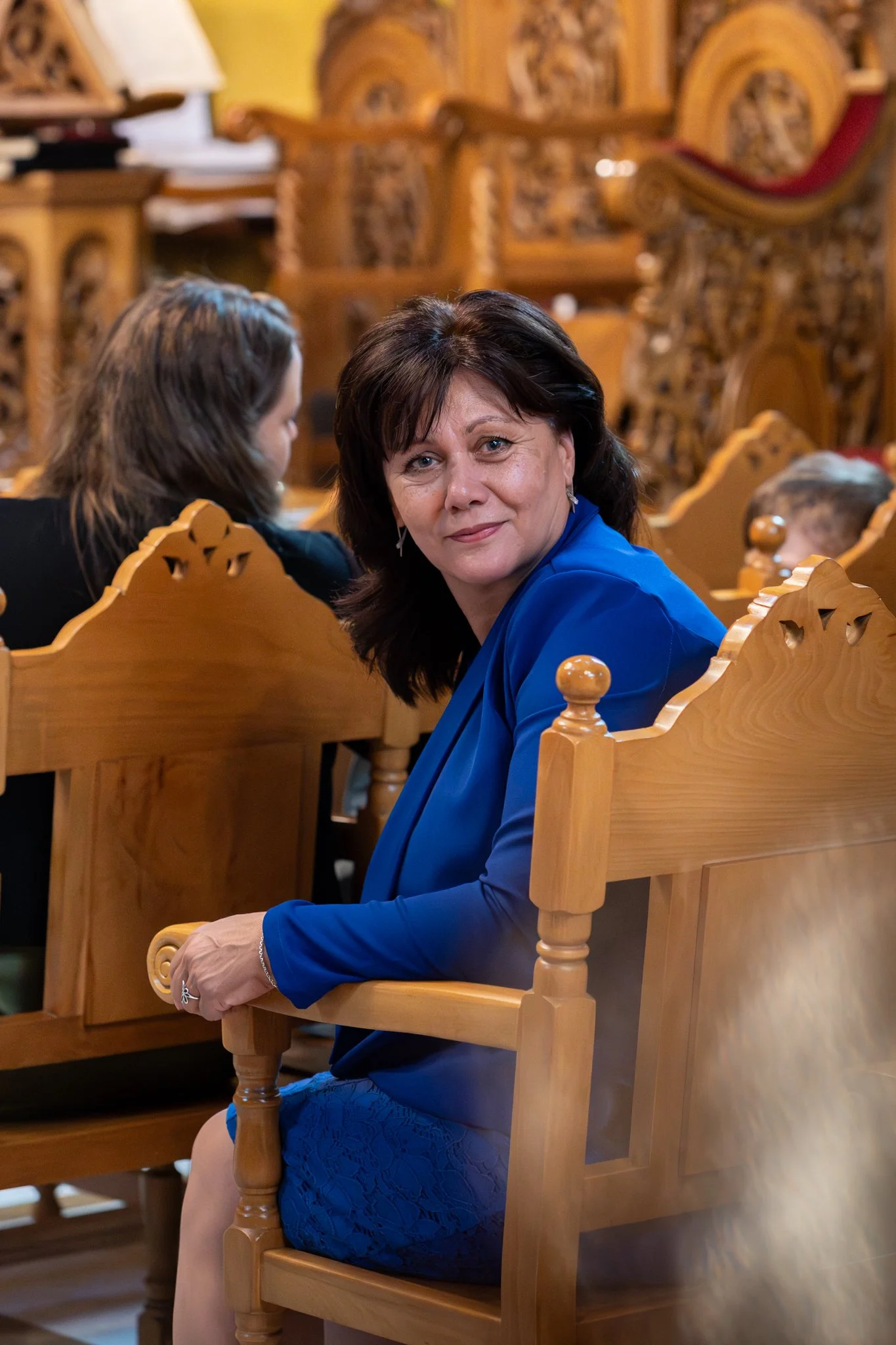 A woman with dark hair wearing a bright blue blazer and matching lace dress sitting on a wooden bench in a room with ornate wood furniture.