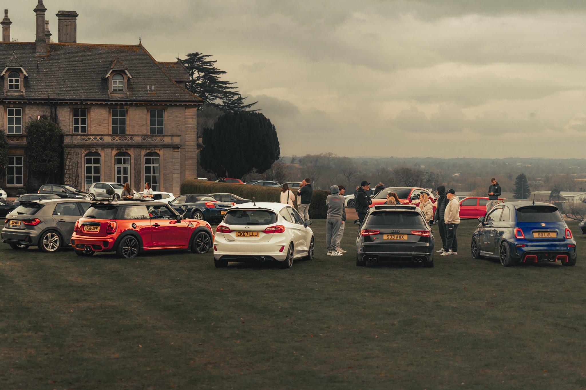 Group of people gathered outdoors near a large historic building with a parking lot filled with various cars, including Audi and Mini Cooper models, under a cloudy sky.