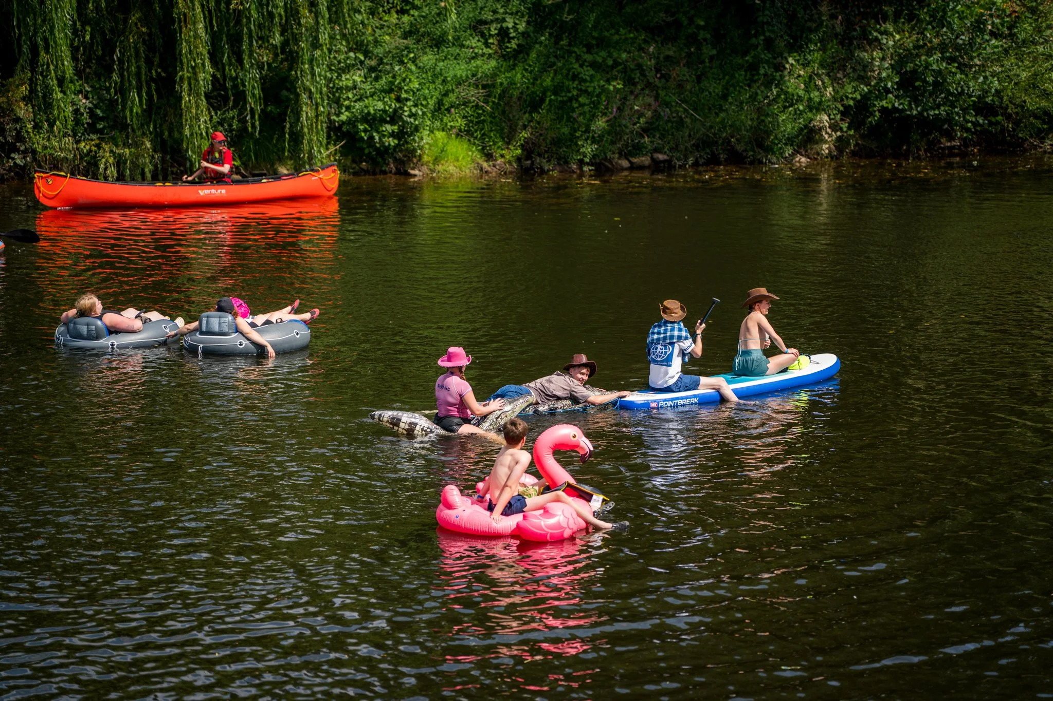 People enjoying a day at the river with various watercraft including a kayak, inflatable tubes, and a paddleboard, surrounded by lush green trees.