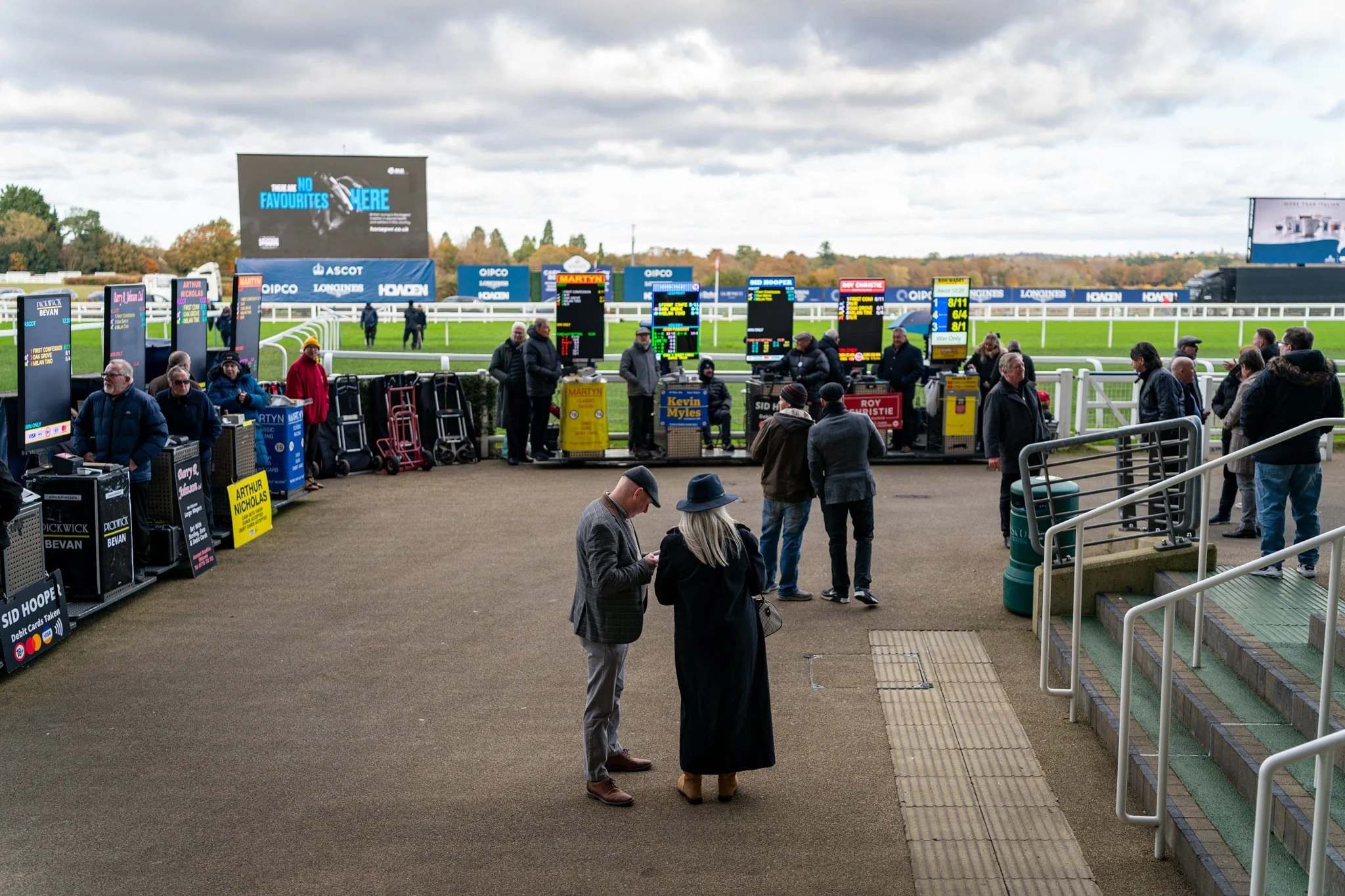 People at a horse racing venue, standing near betting terminals, with horse race odds displayed on electronic boards and a horse racing track in the background under cloudy skies.