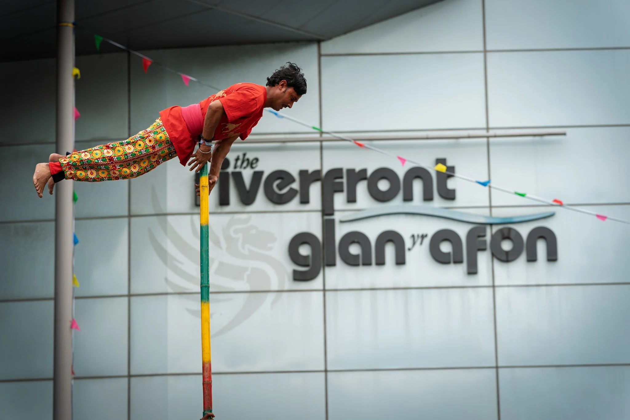A man dressed in colorful, patterned pants and a red shirt is balancing on a tall, multicolored pole during a performance or event. The background features a gray wall with the partially visible text 'the riverfront' and colorful triangular banners h