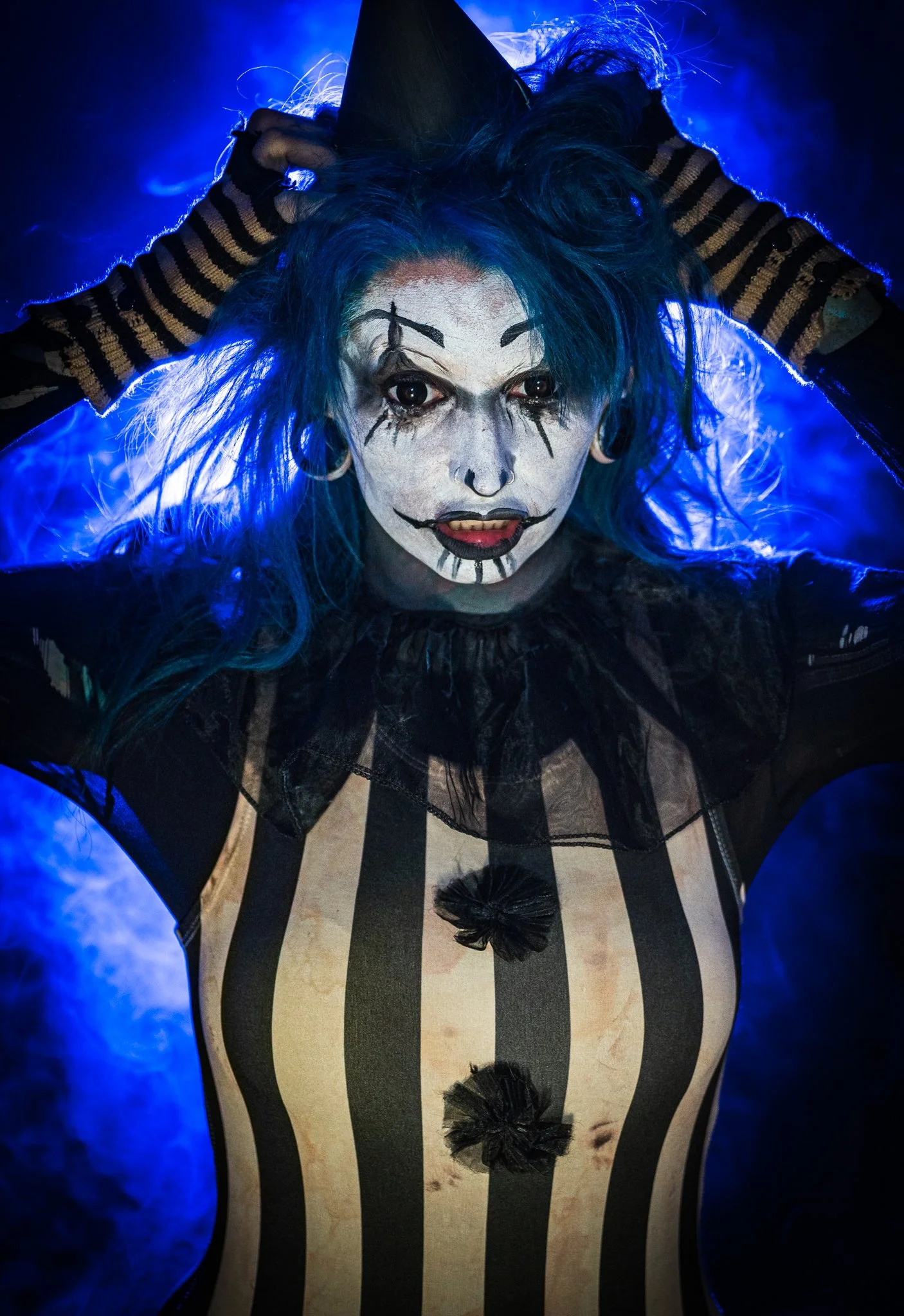 Woman with blue hair, makeup resembling a clown or mime, striped clothing, black tights with pom-pom decorations, striking a pose with hands behind head, dark background with blue lighting.