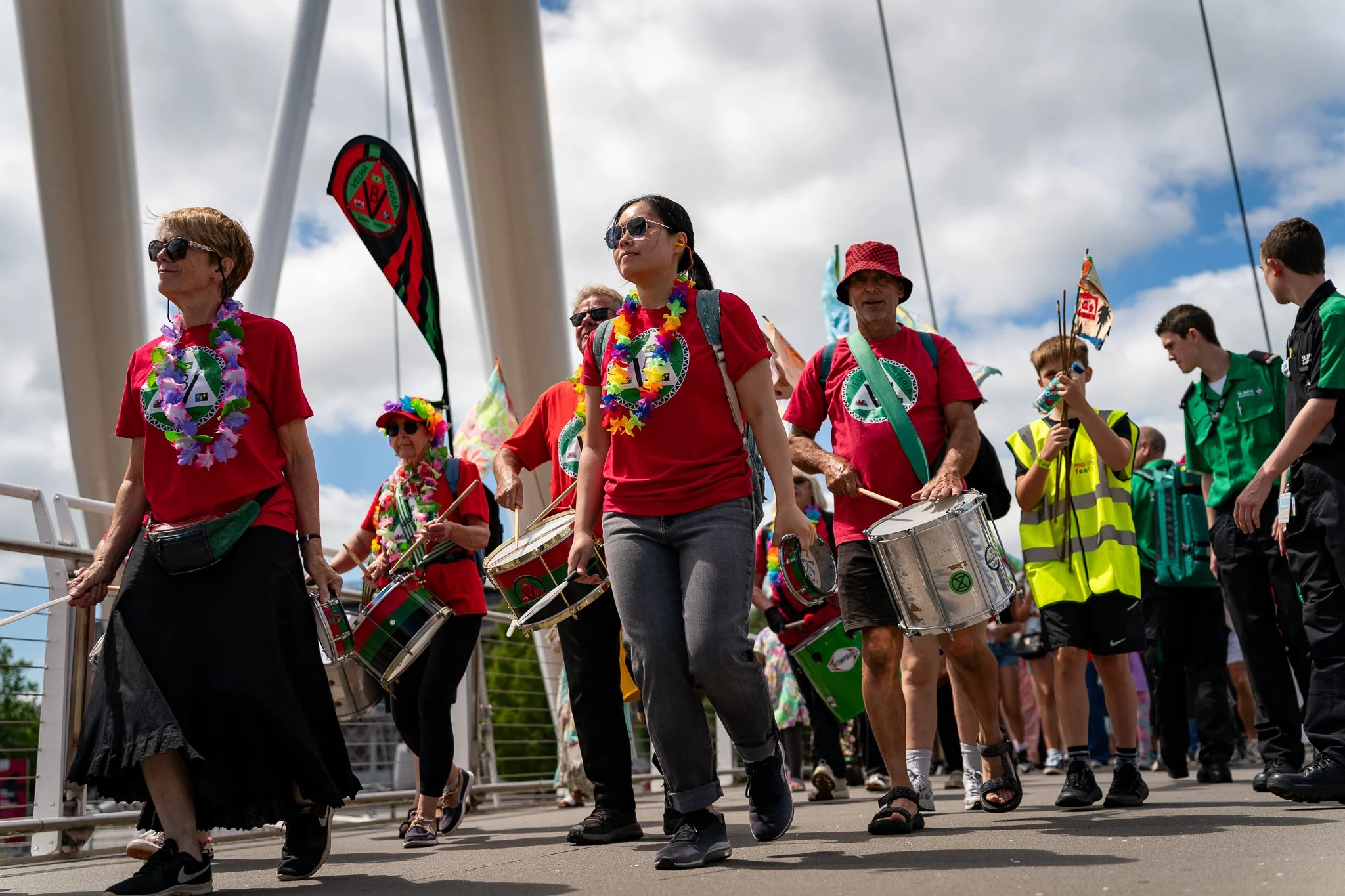 Group of people participating in a parade or celebration, some wearing colorful leis and red T-shirts, playing drums and walking outside under a cloudy sky.