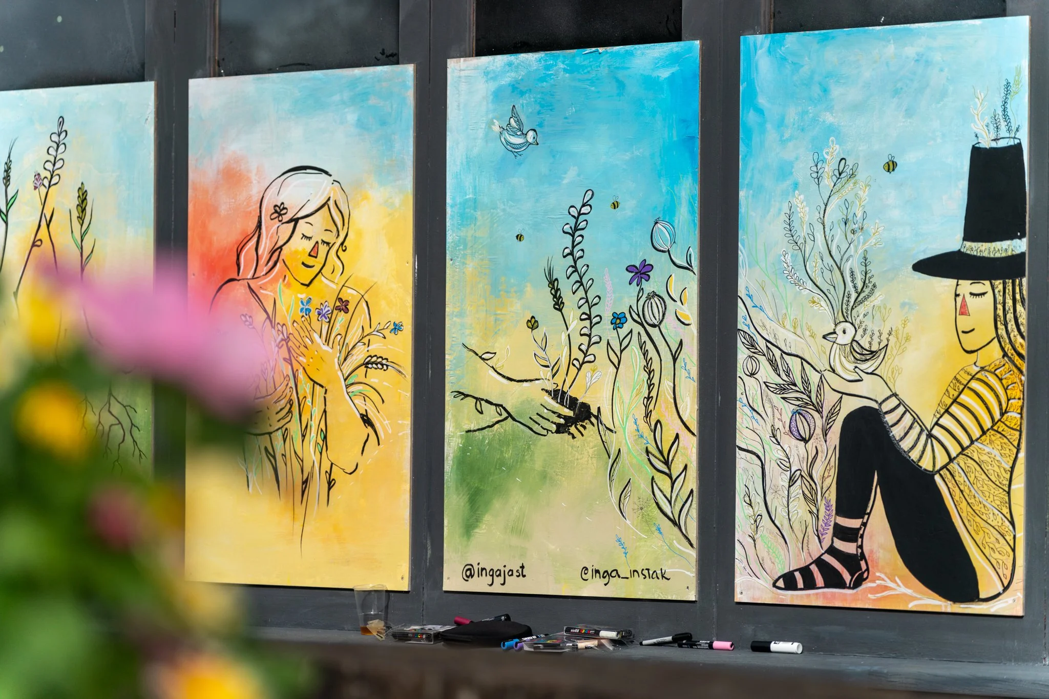 Three colorful paintings with black outlines of women, plants, bees, and birds, displayed on a dark wall. Markers are placed on a ledge below the paintings, with some masking tape visible at the bottom.