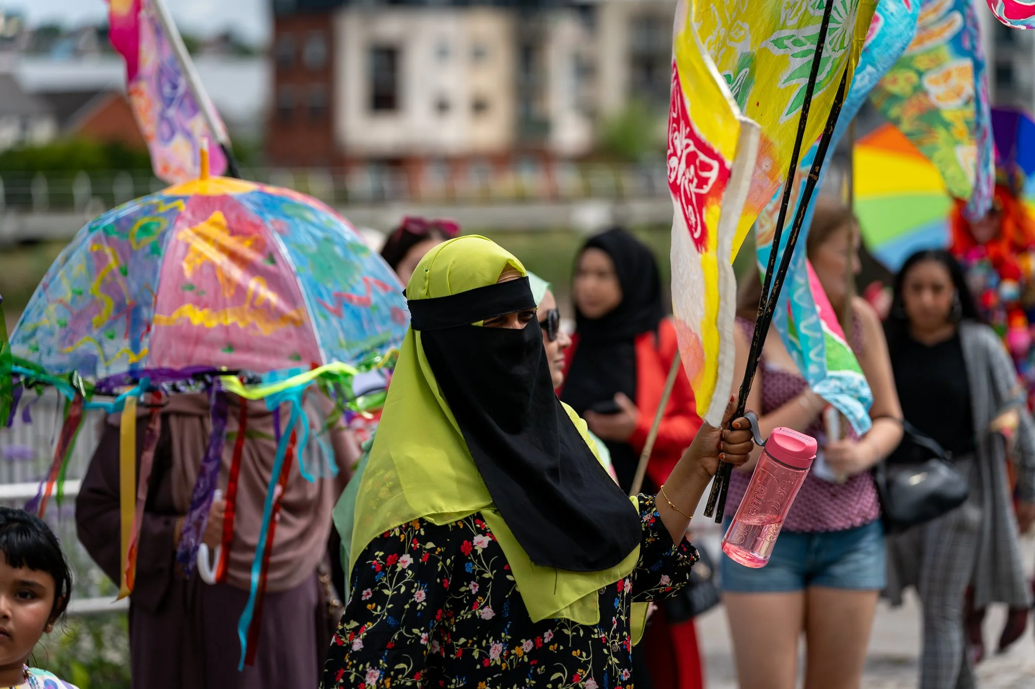 Group of people at an outdoor event, some holding colorful umbrellas and flags, one woman wearing a bright green hijab and black face covering, holding a pink water bottle.