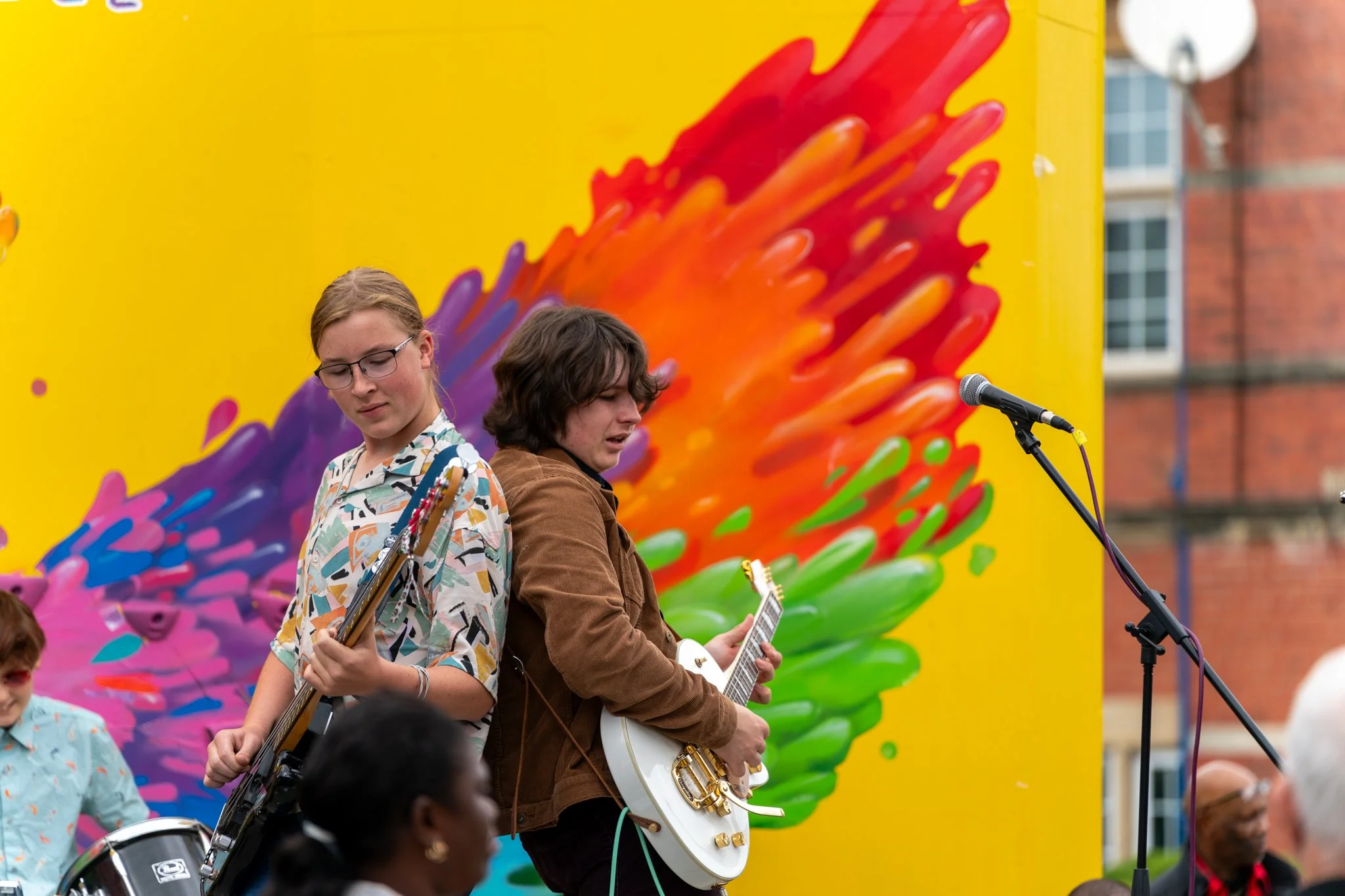 Two young men with guitars performing on stage at an outdoor event, with a bright yellow background featuring colorful abstract art.