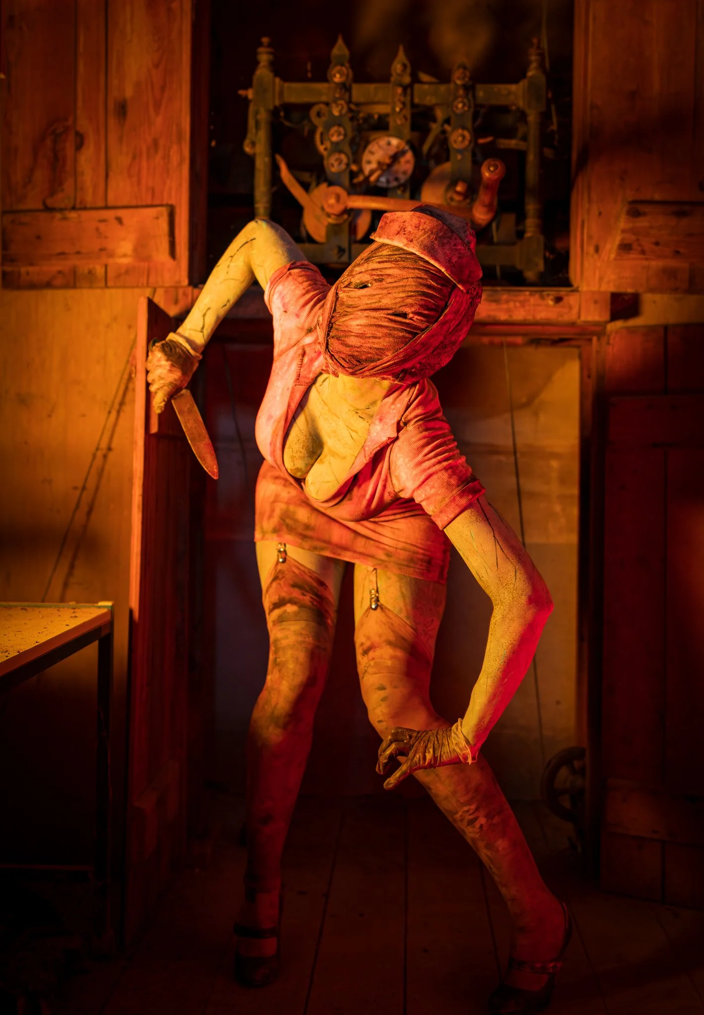 A person dressed in a humanoid costume with a distorted face in a room with wooden walls and furniture, standing with one leg bent and holding a knife.
