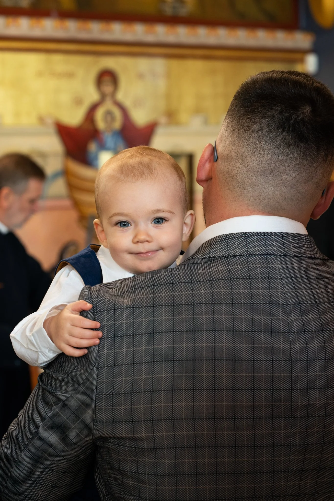 A young boy with light skin and blue eyes looking directly at the camera while being held by a man with dark hair and a checkered suit. In the background, there is religious iconography on a wall, suggesting the location is a church or similar settin