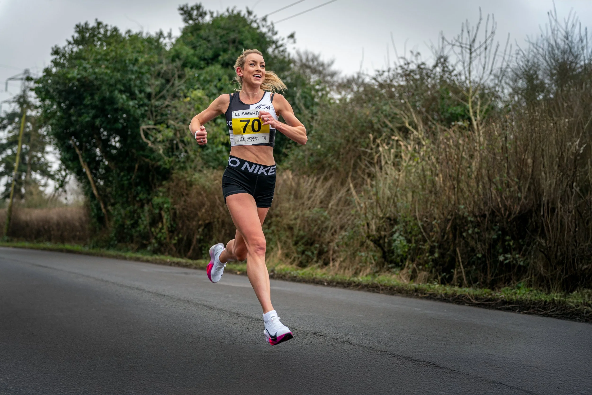Female runner in black athletic gear running outdoors on a paved road surrounded by trees and shrubs on a cloudy day.