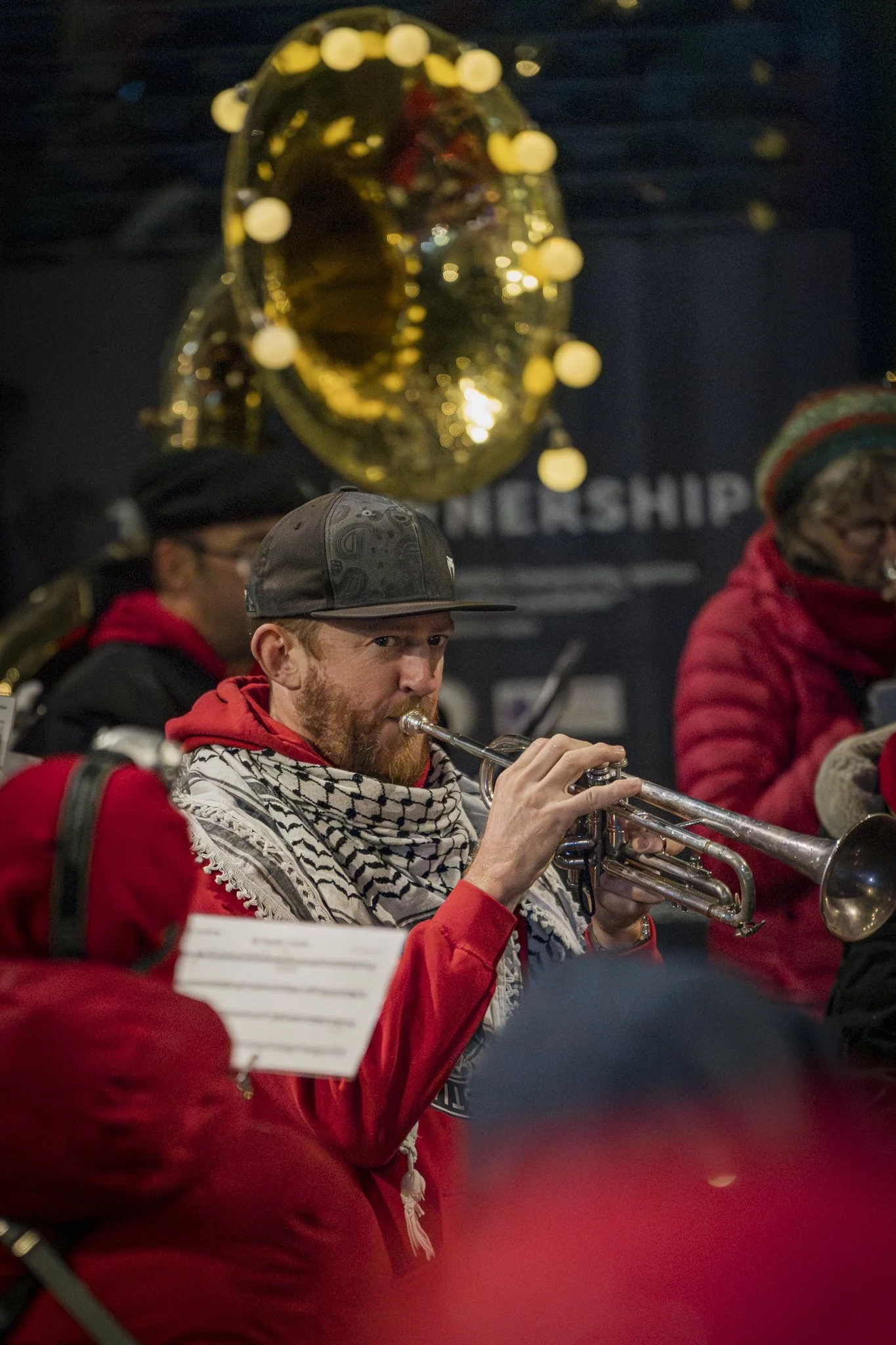A man with a beard wearing a black cap, red hoodie, and a patterned scarf playing a trumpet. There are other musicians around him, and a large shiny golden medal or trophy is hanging above them.