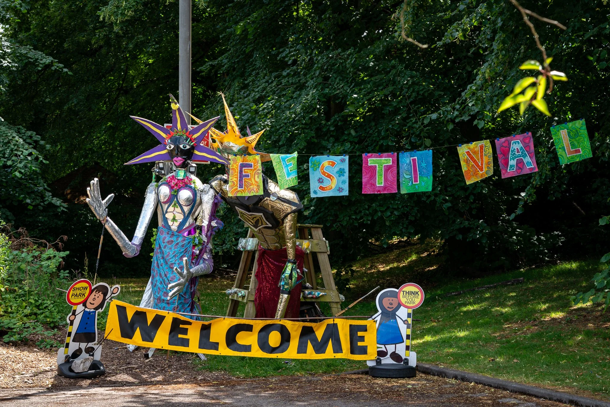 Colorful outdoor display with a large puppet or statue representing a character with a star-shaped hat, surrounded by signs saying 'Welcome', 'Festivals' and other messages, set in a wooded park area.