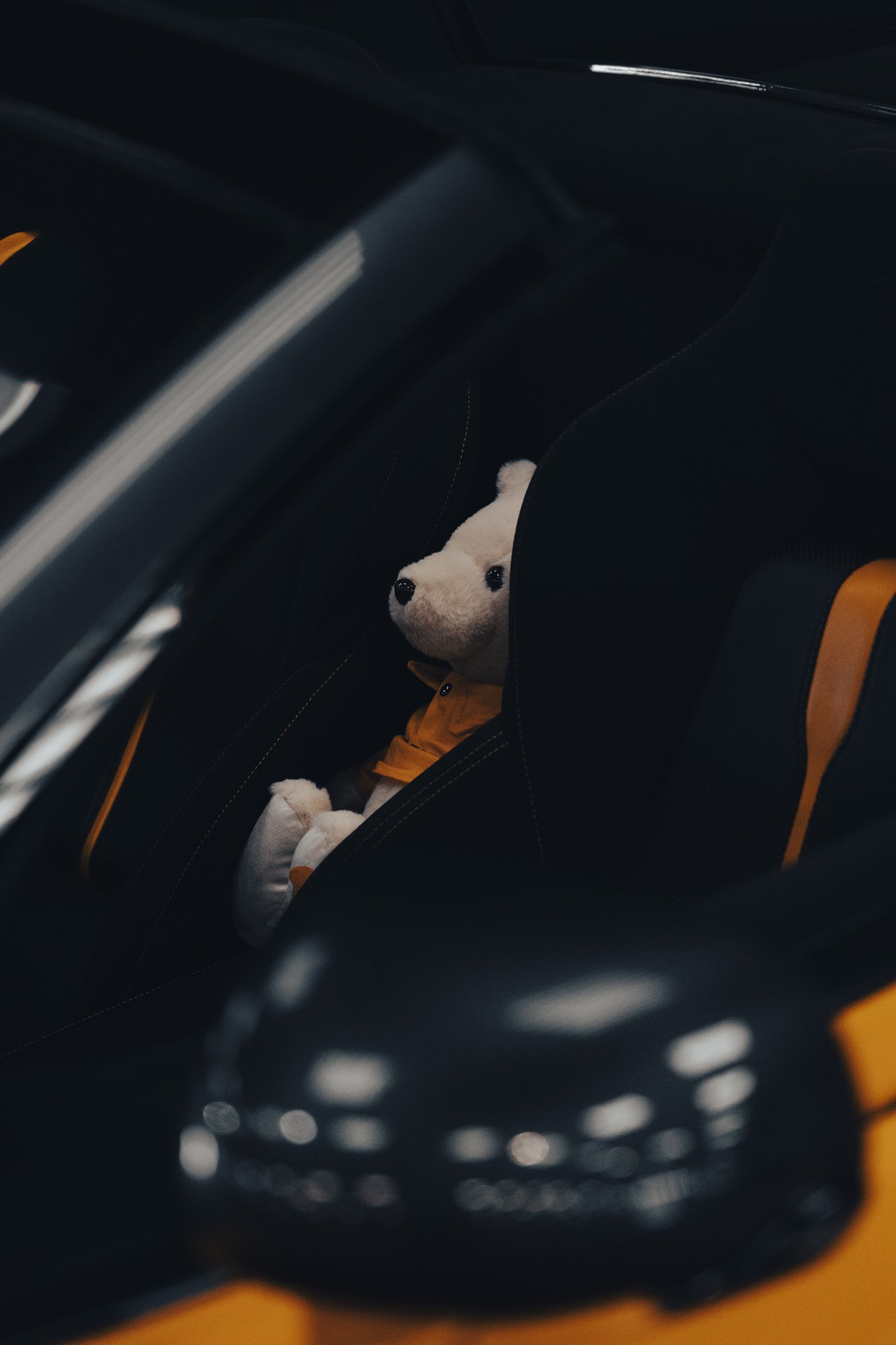 A plush teddy bear wearing a yellow jacket and white pants sitting in the backseat of a car, partially visible through the car's headrest.