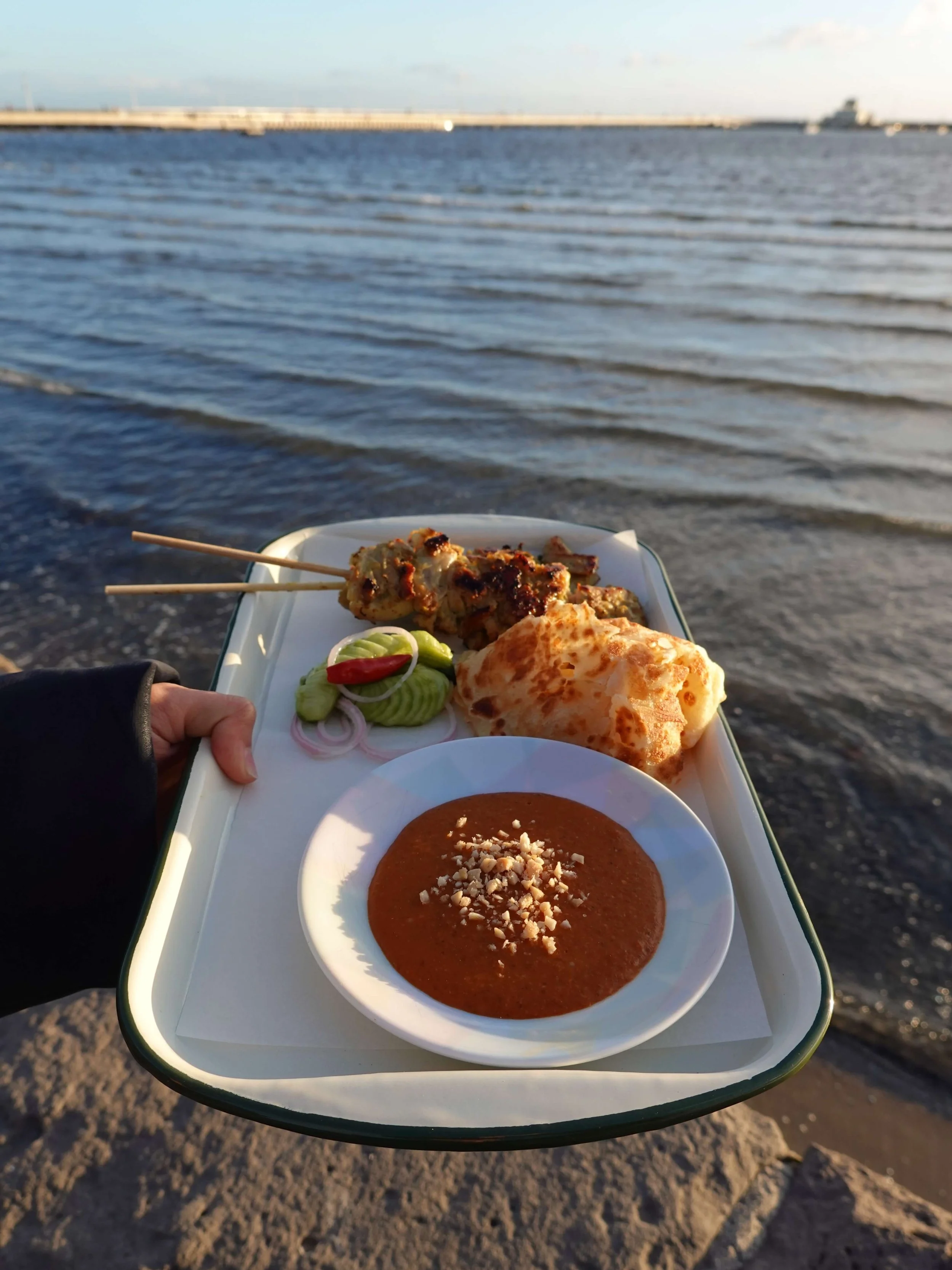A tray of food held by a person near the shoreline. The tray contains grilled skewers, a small salad with cucumber, onion, and red pepper, a piece of bread, and a bowl of brown sauce topped with crushed nuts. The ocean and a distant pier or breakwater are visible in the background.