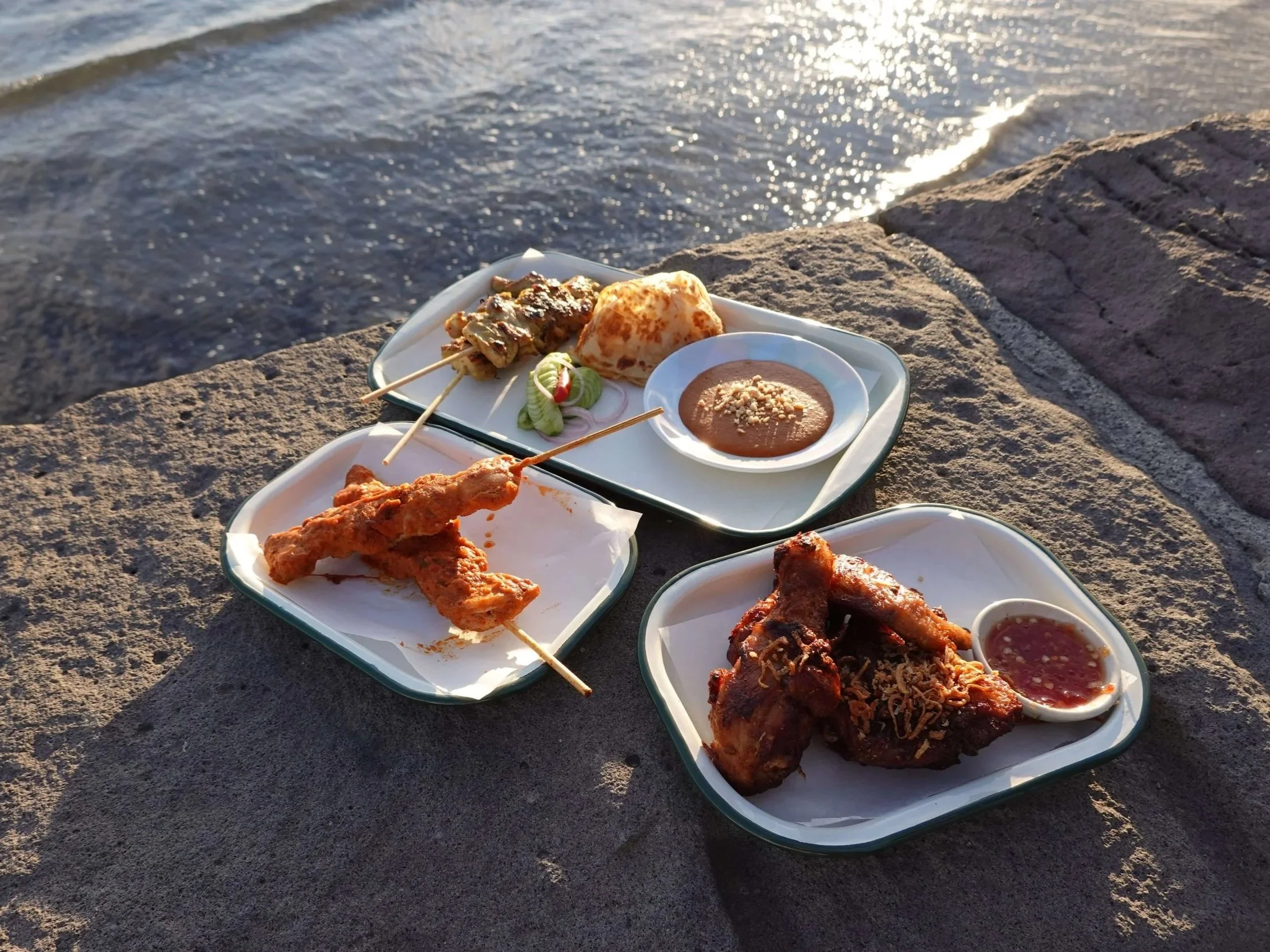 A tray of food including skewered meat, a bowl of sauce, and fried chicken pieces, placed on a rocky surface near a body of water during sunset.