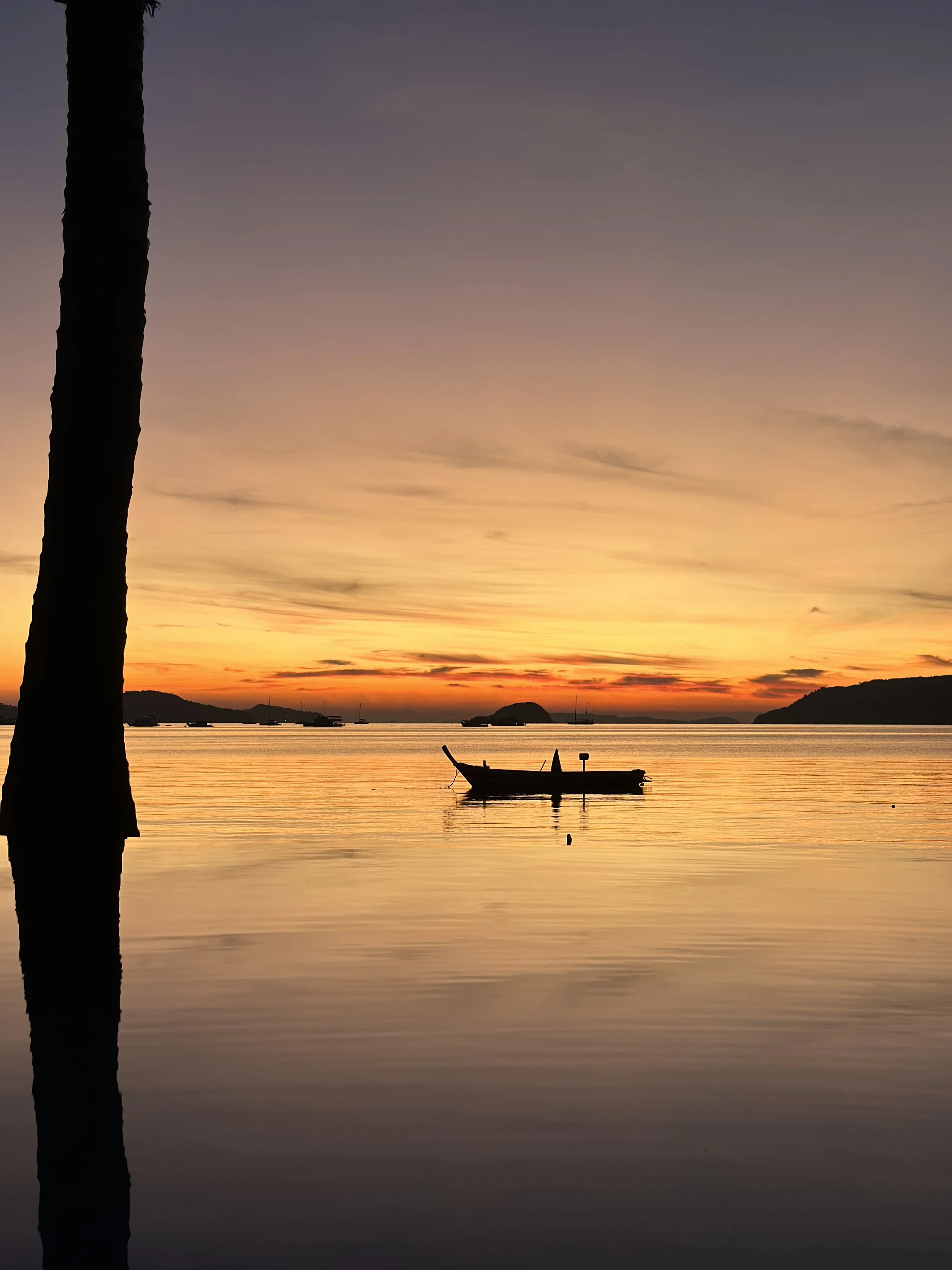 A silhouette of a palm tree trunk on the left with a calm body of water and a small boat in the foreground. The sunset in the background features a colorful sky with shades of purple, orange, and yellow, and distant islands or landforms on the horizon.