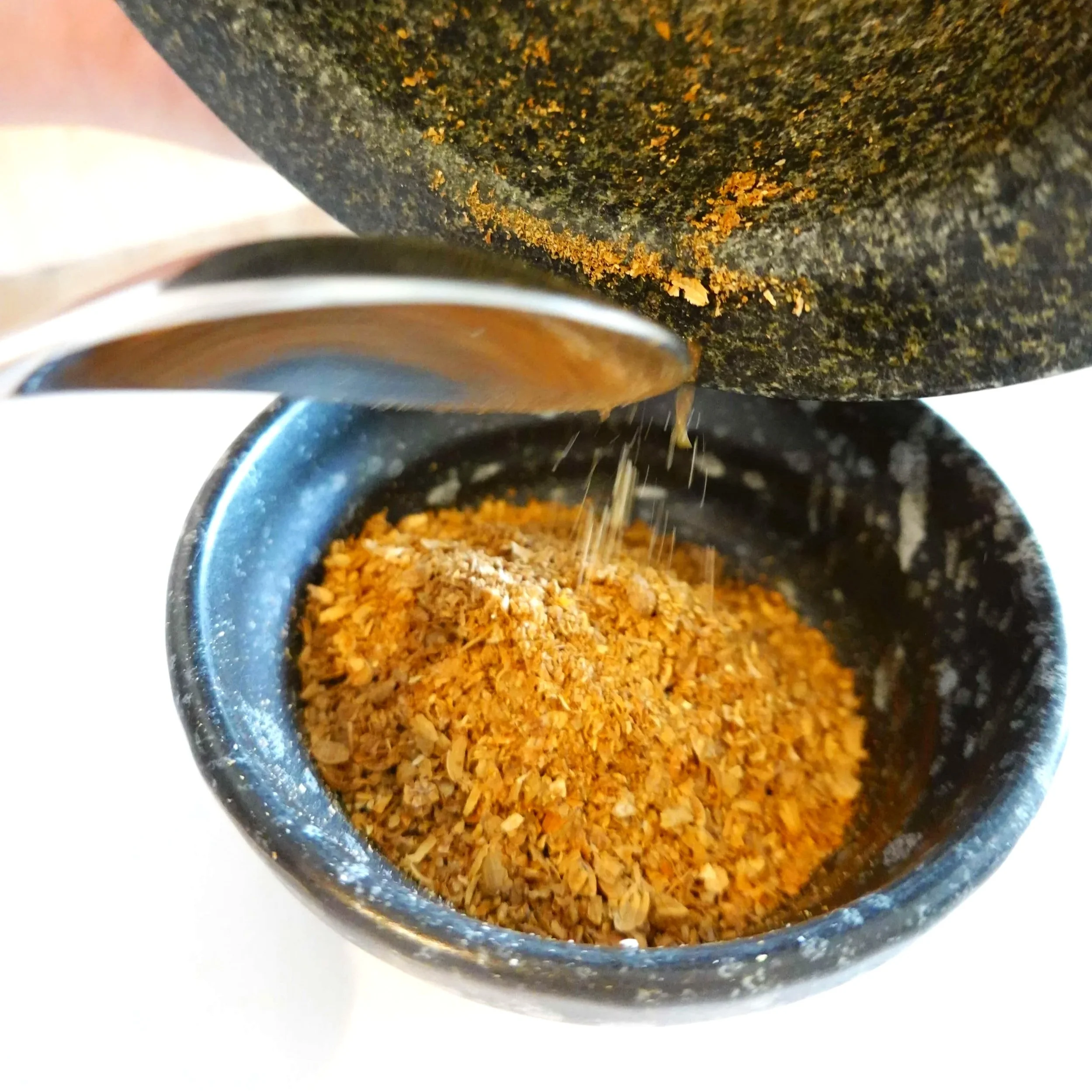 A close-up of a blue speckled ceramic cup filled with freshly grinded herbs or spices, with a silver spoon resting on the edge of the cup.