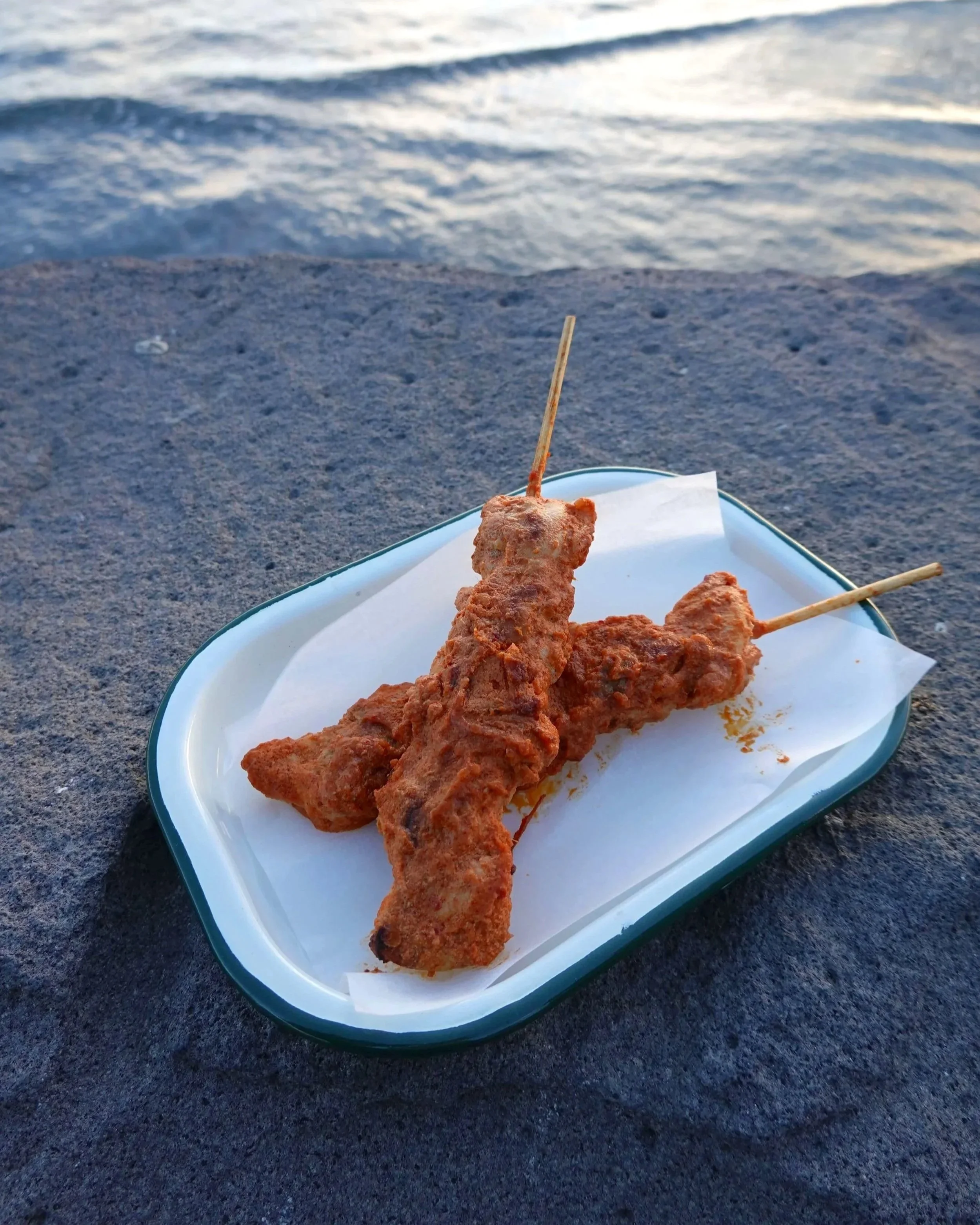Freshly grilled chicken skewered marinated in coconut kolae curry sauce on a white tray, placed on a dark surface with a background of water.