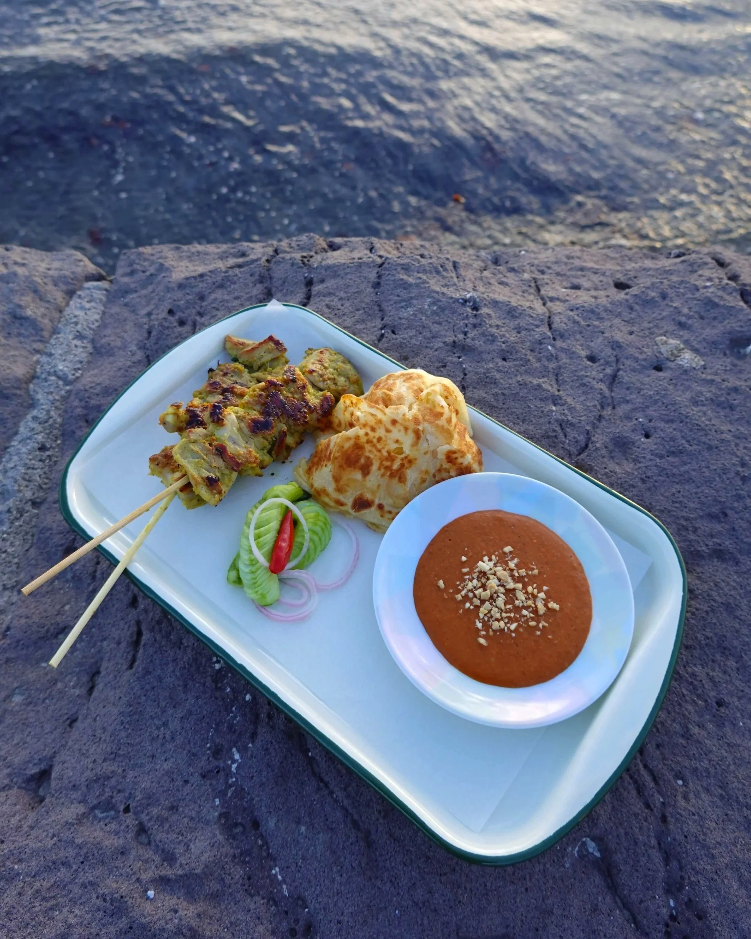 A white tray with green border containing grilled fresh roti, green pickles with chilli, and a bowl of satay peanut sauce garnished with chopped roasted nuts on a rocky surface by the water.