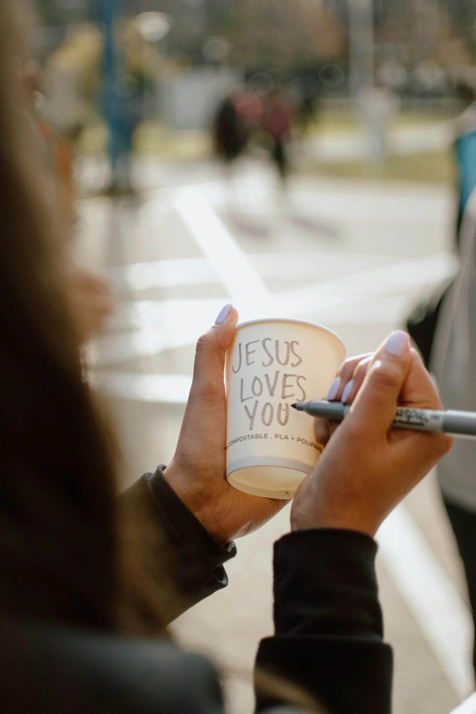A person holding a paper cup with the handwritten message "JESUS LOVES YOU" and signing it with a black marker. The scene takes place outdoors with people walking in the background.