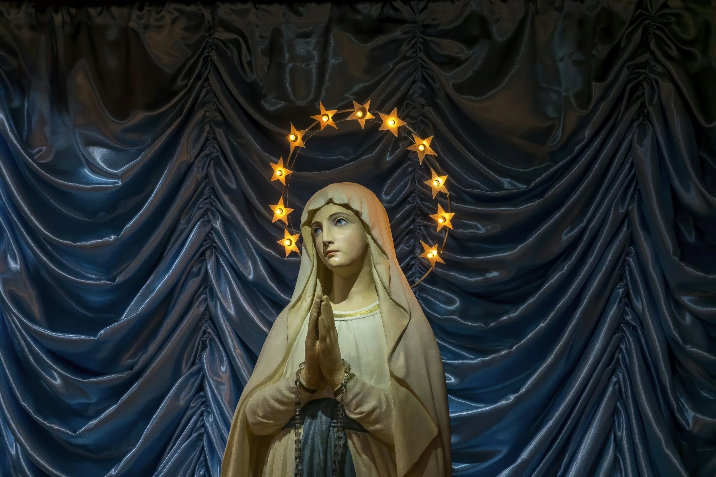 Statue of Virgin Mary with hands in prayer, standing in front of a dark, shiny, pleated fabric backdrop. A halo of illuminated star-shaped lights surrounds her head.