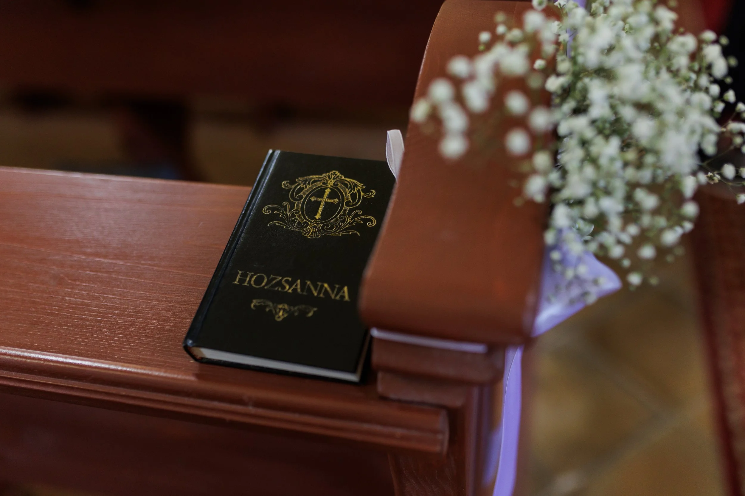 A close-up photo of a prayer book and a bouquet of white flowers placed on a wooden surface, with a pinkish-brown background.