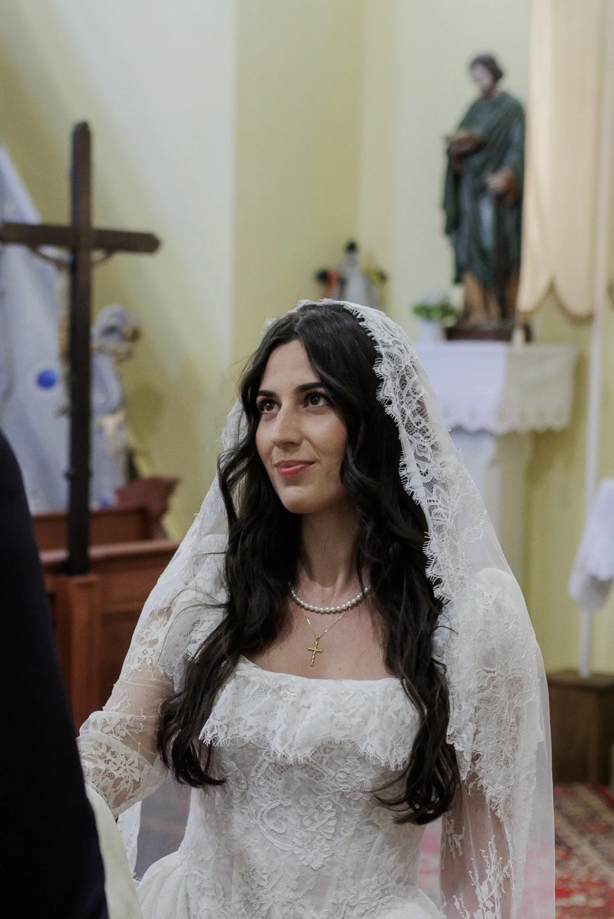 A bride with long, dark, wavy hair wearing a white lace wedding dress and a pearl necklace with a gold cross pendant, standing inside a church with a religious statue and cross in the background.