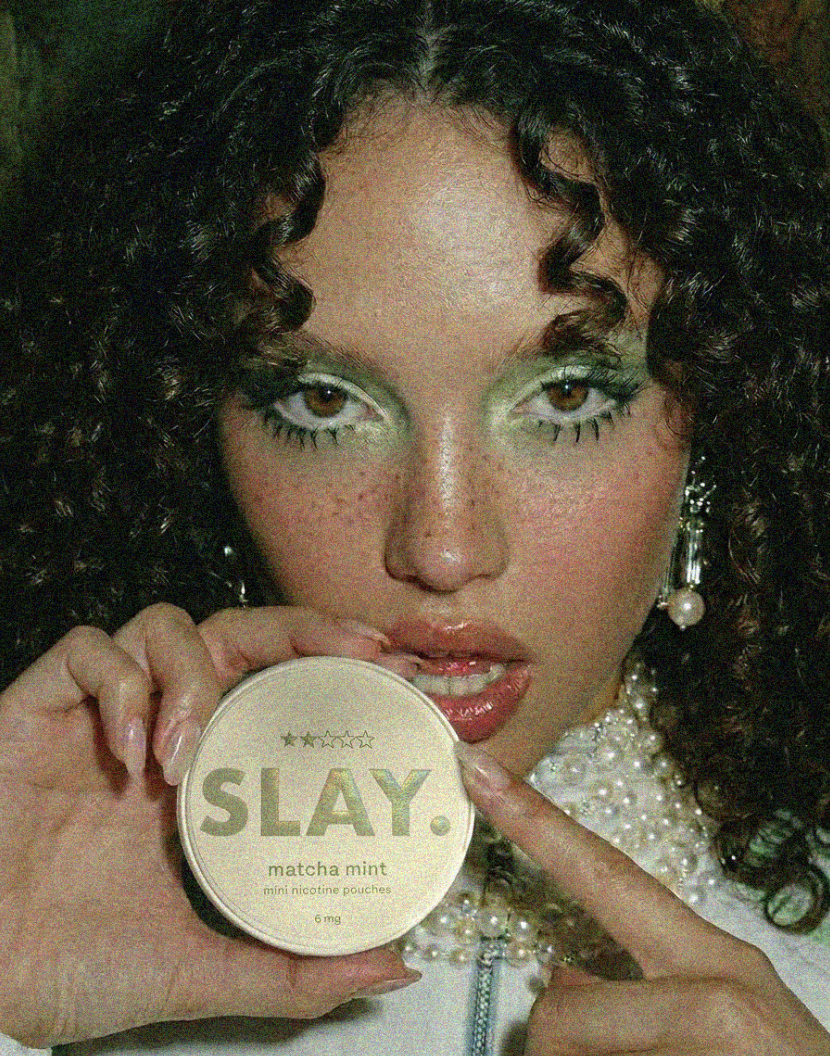 A woman with curly hair and makeup holding a round container labeled 'SLAY. matcha mint mini nicotine pouches' near her lips with her finger.