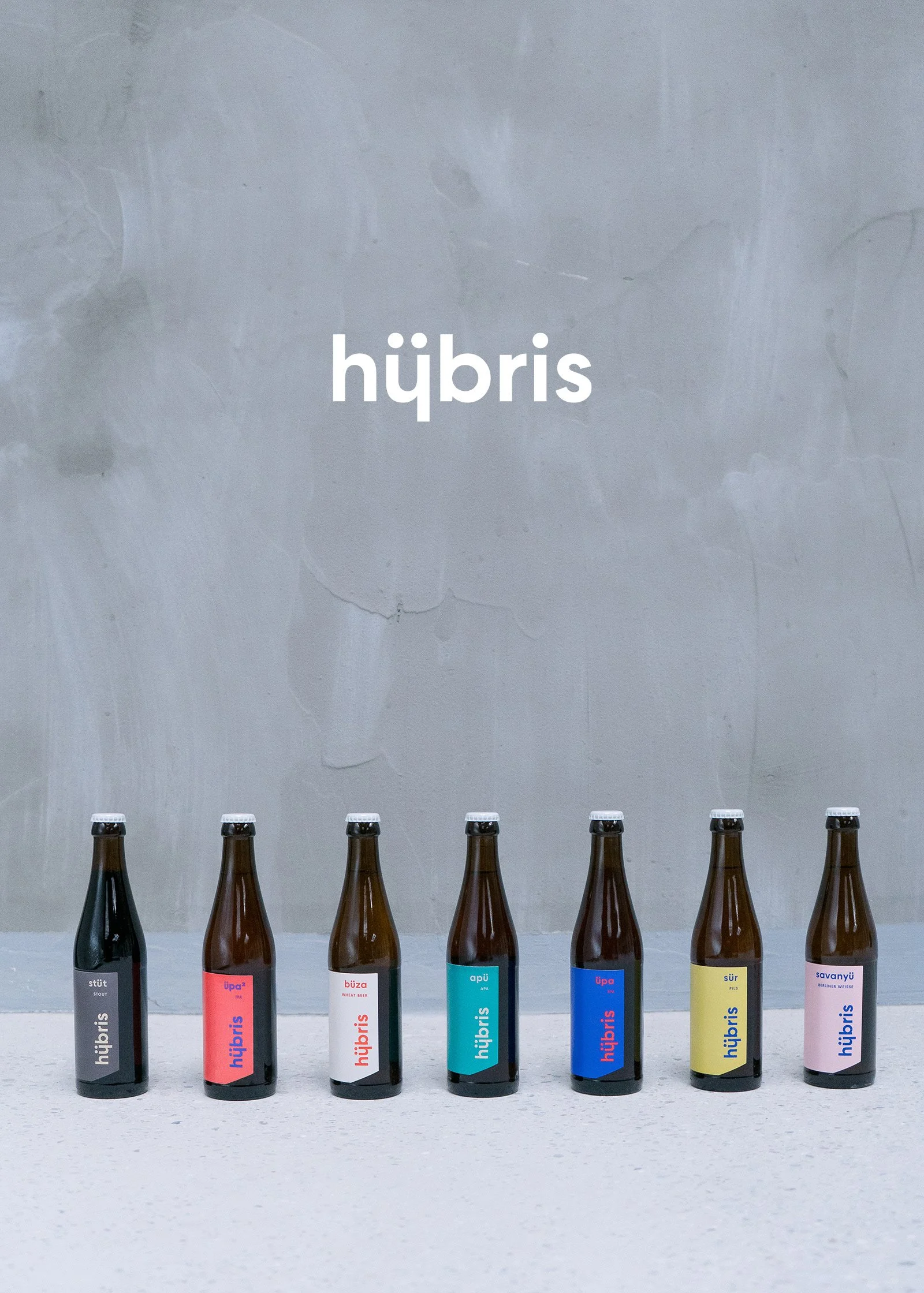 Six bottles of bubbly drinks with colorful labels, standing in a row on a white table, with a gray wall behind them that has the word 'hübris' written on it.