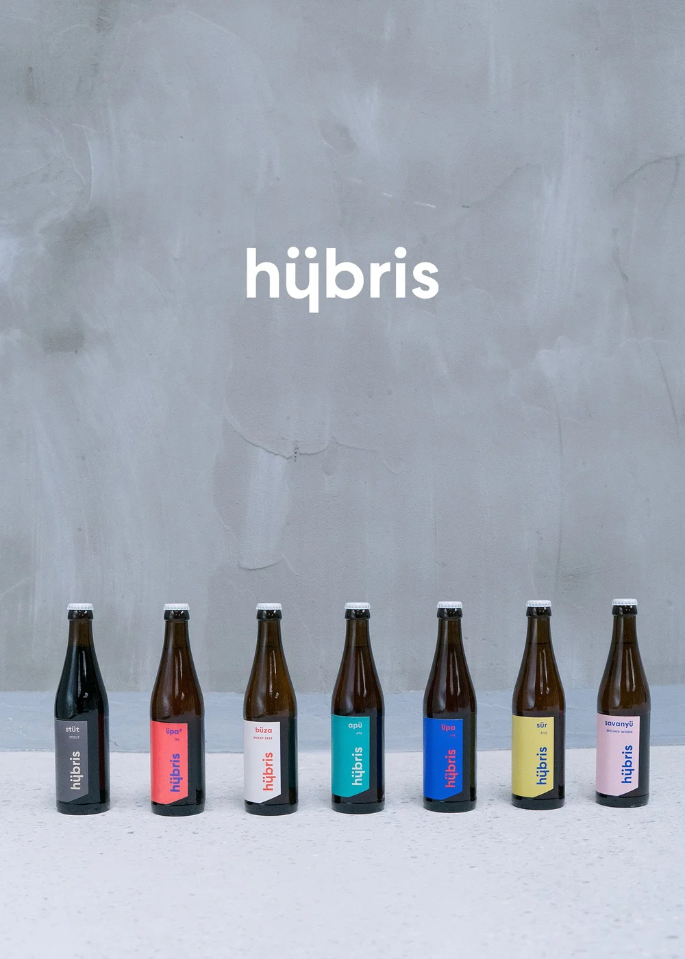 Six bottles of bubbly drinks with colorful labels, standing in a row on a white table, with a gray wall behind them that has the word 'hübris' written on it.