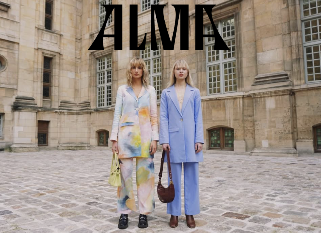 Two women standing outdoors in front of a historic stone building with multiple windows. One woman wears a colorful tie-dye blazer and pants, and the other wears a light blue blazer and pants. The word 'ALMA' is displayed above them.