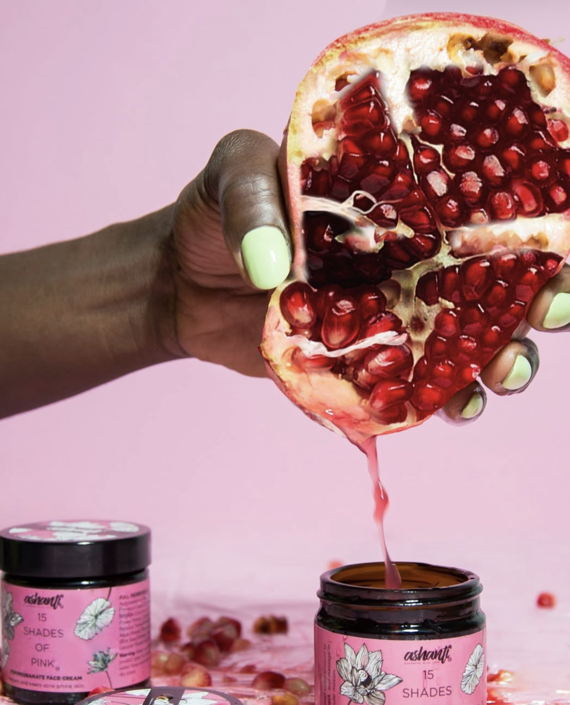 A person holding a halved pomegranate above a jar of pink face cream with pink background.