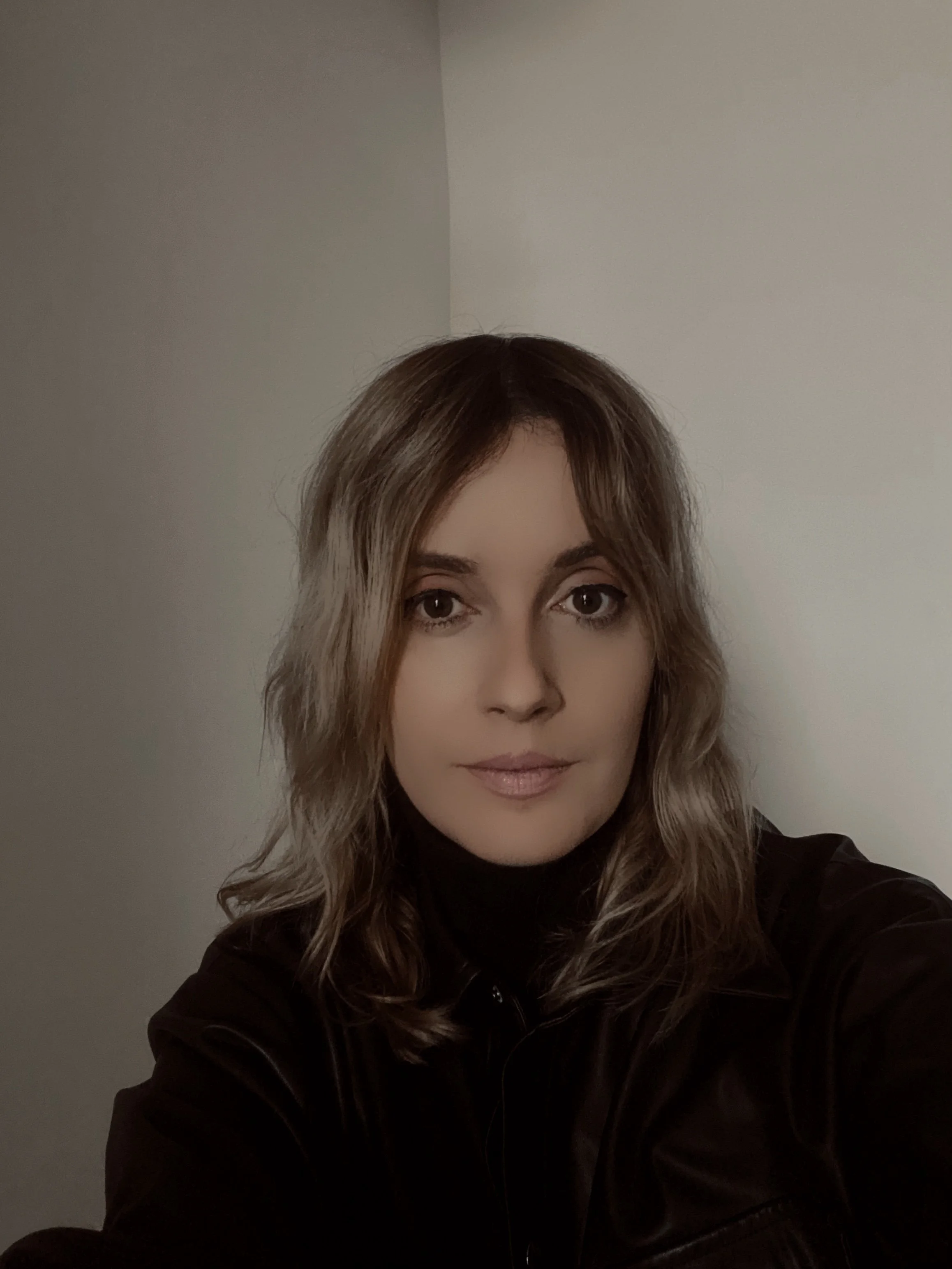 A woman with wavy, shoulder-length hair taking a selfie indoors against a plain, light-colored wall. She is wearing a dark top and has a neutral expression.