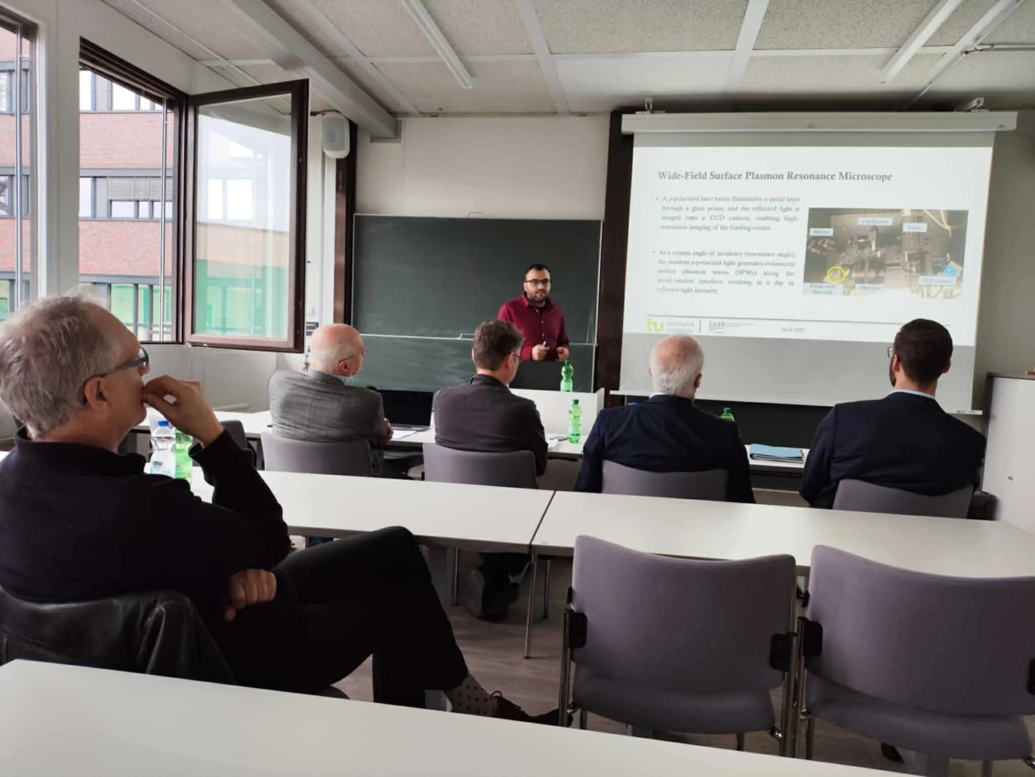 A man presenting in a classroom with a projector screen displaying scientific information. Audience members are seated, listening attentively.