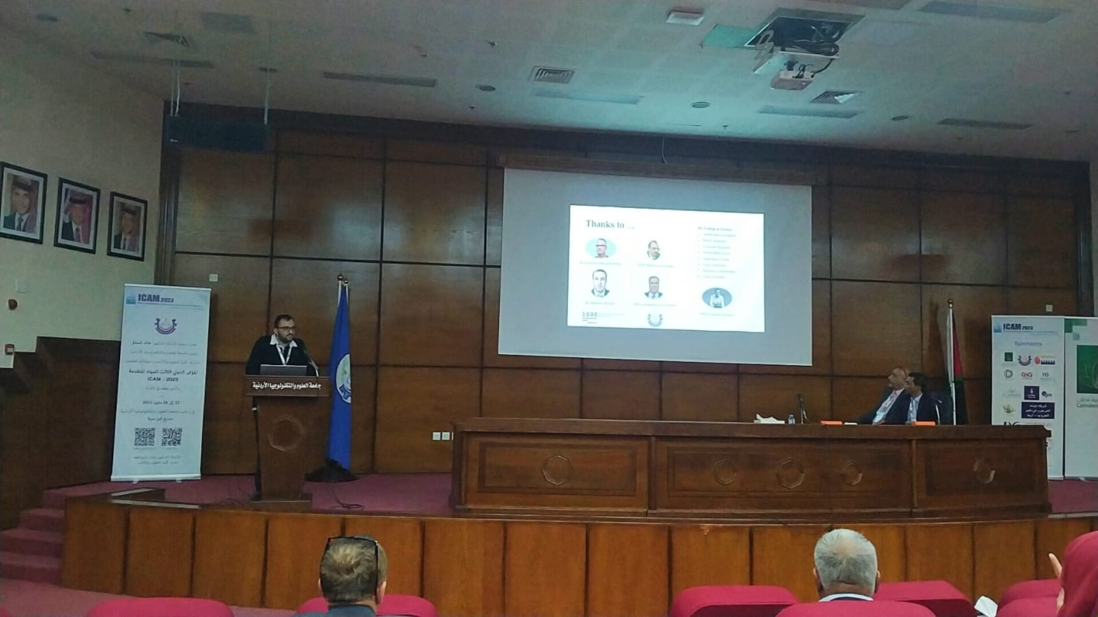 A conference room with a speaker at a podium, a large screen displaying a presentation, and two attendees seated at a long desk. The room has portraits on the wall, flags, and banners with conference information.