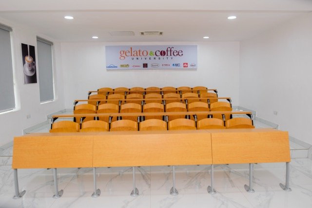 Empty classroom with orange chairs arranged in four rows, wooden desk at the front, white walls, a banner on the wall that reads 'gelato & coffee university,' and framed photo of coffee on the left wall.