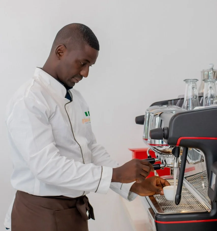A chef in a white uniform prepares coffee using an espresso machine in a kitchen or café.